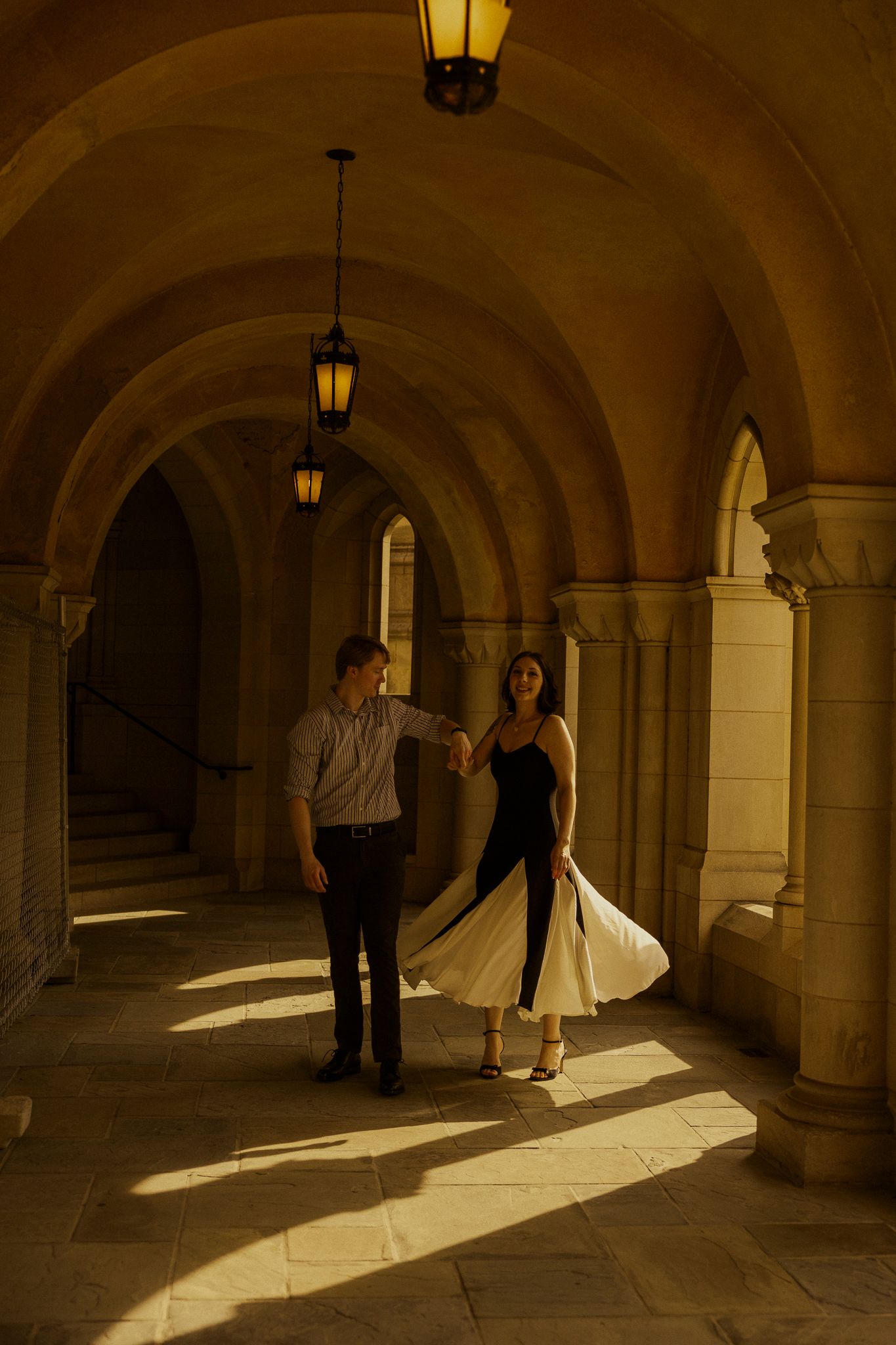 Couple dancing in golden-lit arched stone hallway, woman in flowing white and black dress, warm lantern light casting dramatic shadows.