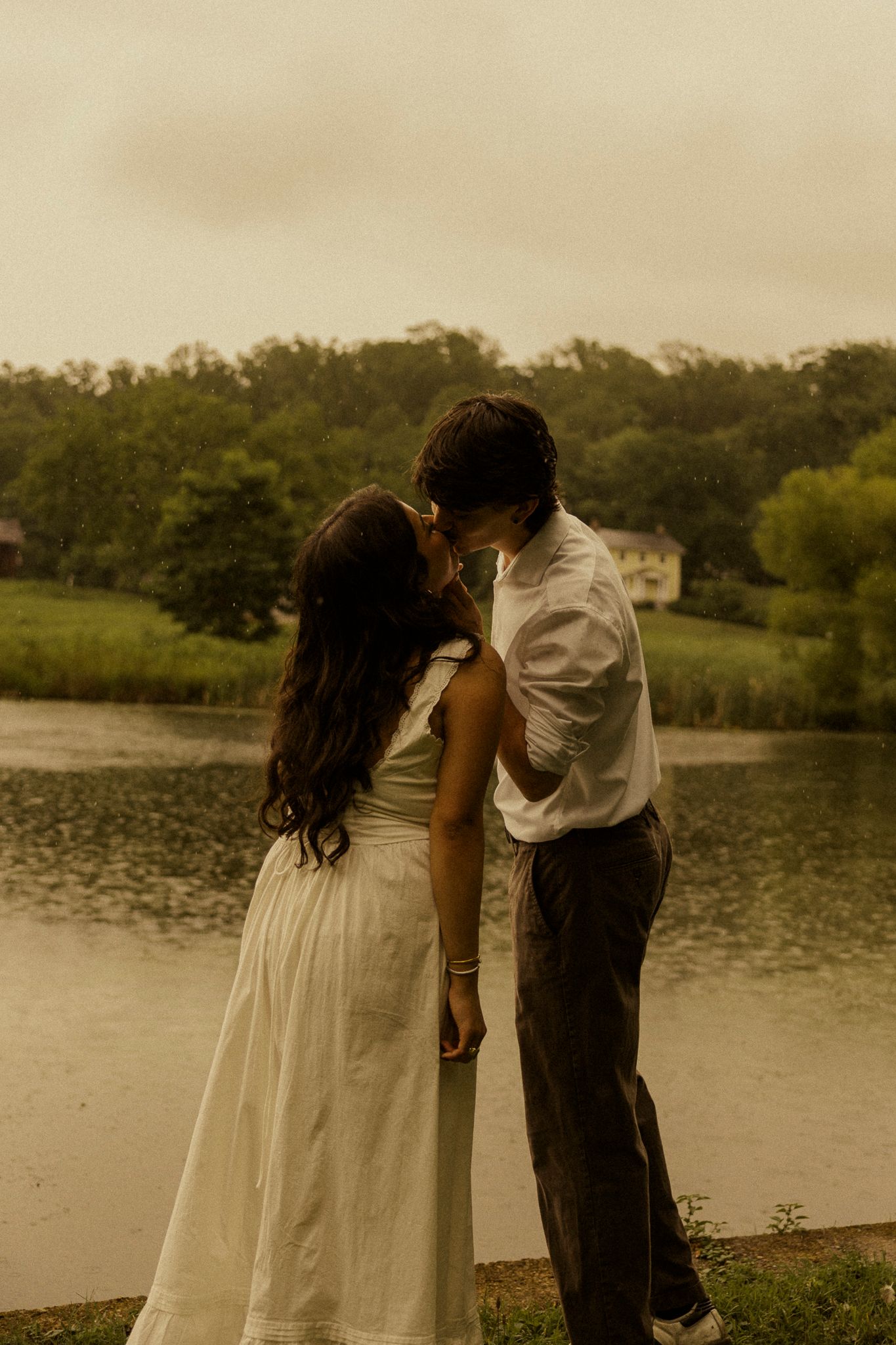 A couple shares a romantic moment by a serene lake, dressed in white, surrounded by lush greenery in soft golden light.