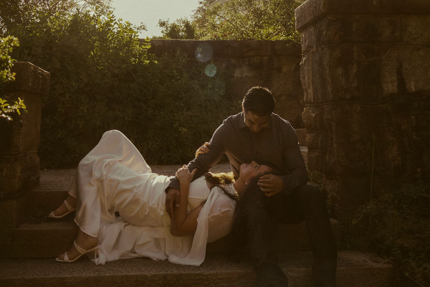 Couple sharing intimate moment on stone steps in golden hour light, bride in white gown resting on groom.