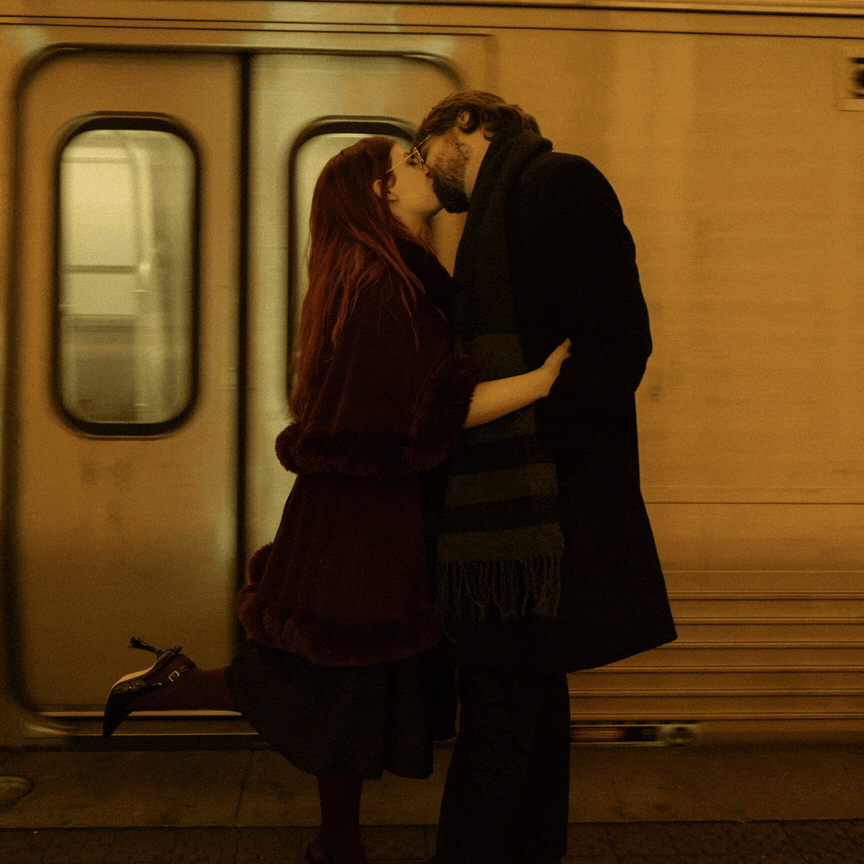 A couple embraces and kisses on a subway platform in front of a train, creating a romantic silhouette against warm lighting.