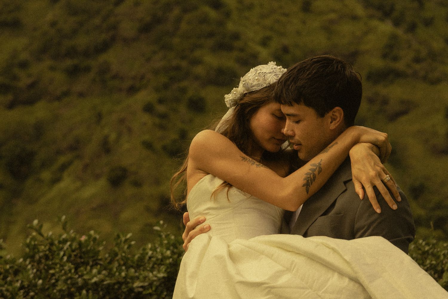 A bride in a white dress and groom in a suit embrace tenderly against a lush green hillside backdrop.