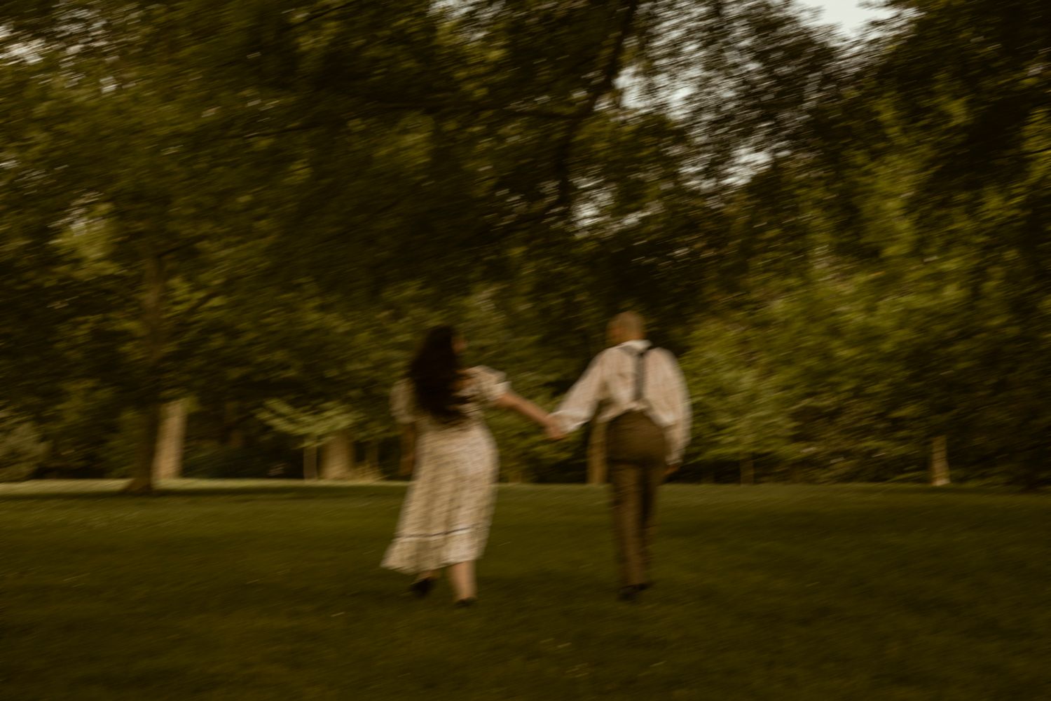 Couple holding hands running through a lush green park in vintage clothing.