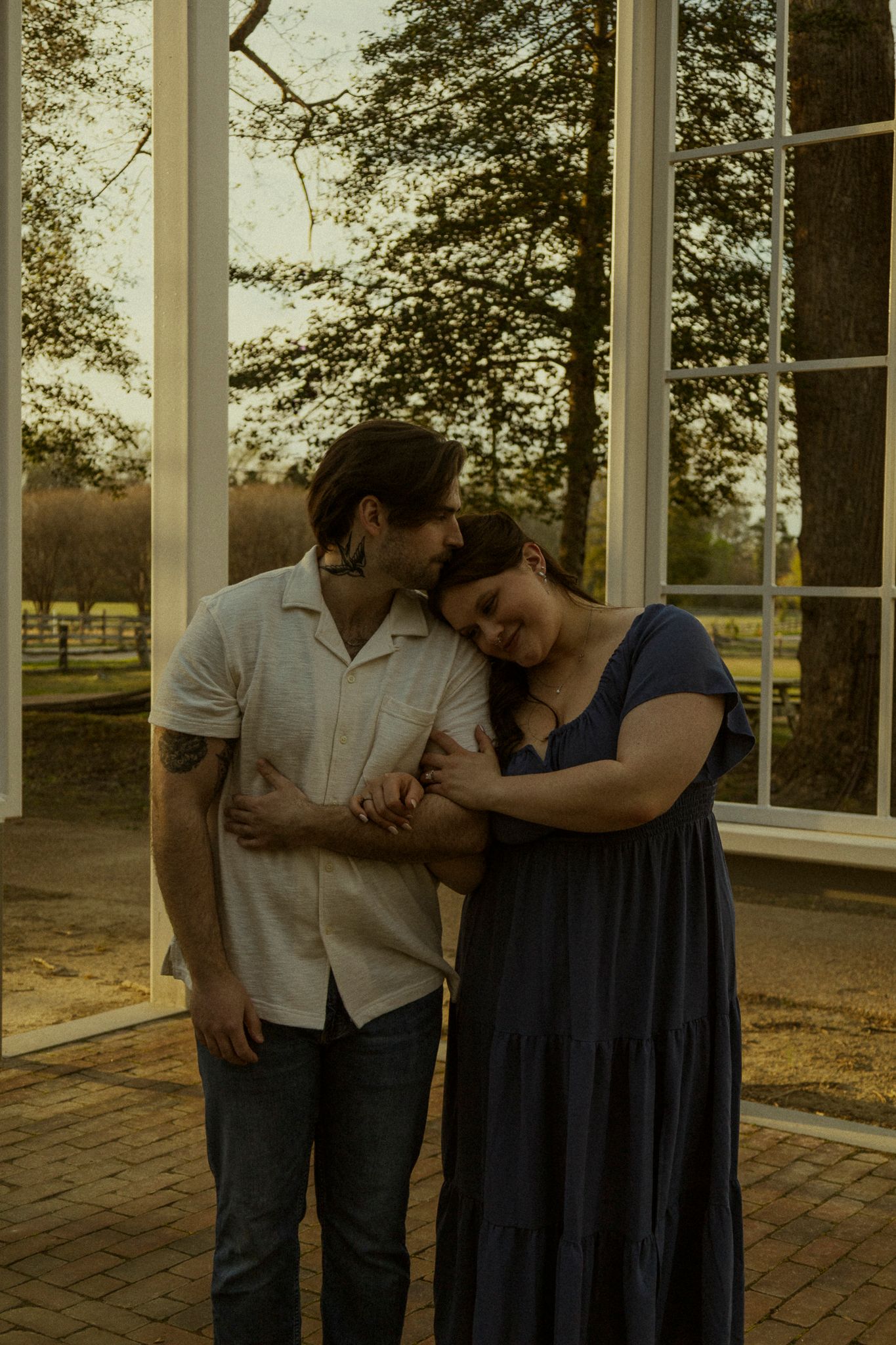 Couple sharing a tender moment embracing near large windows with golden hour light at an outdoor venue.