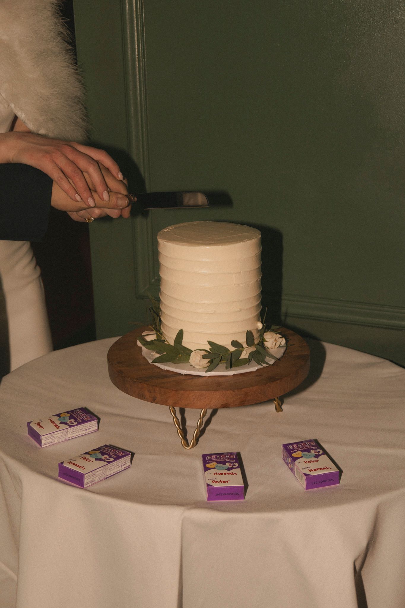 Couple cutting a simple white textured wedding cake decorated with greenery on a wooden stand at reception.