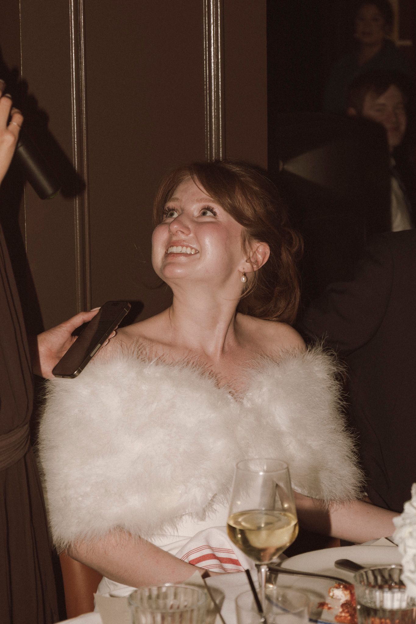 Smiling bride in white off-shoulder gown with fluffy wrap shawl seated at wedding reception table.