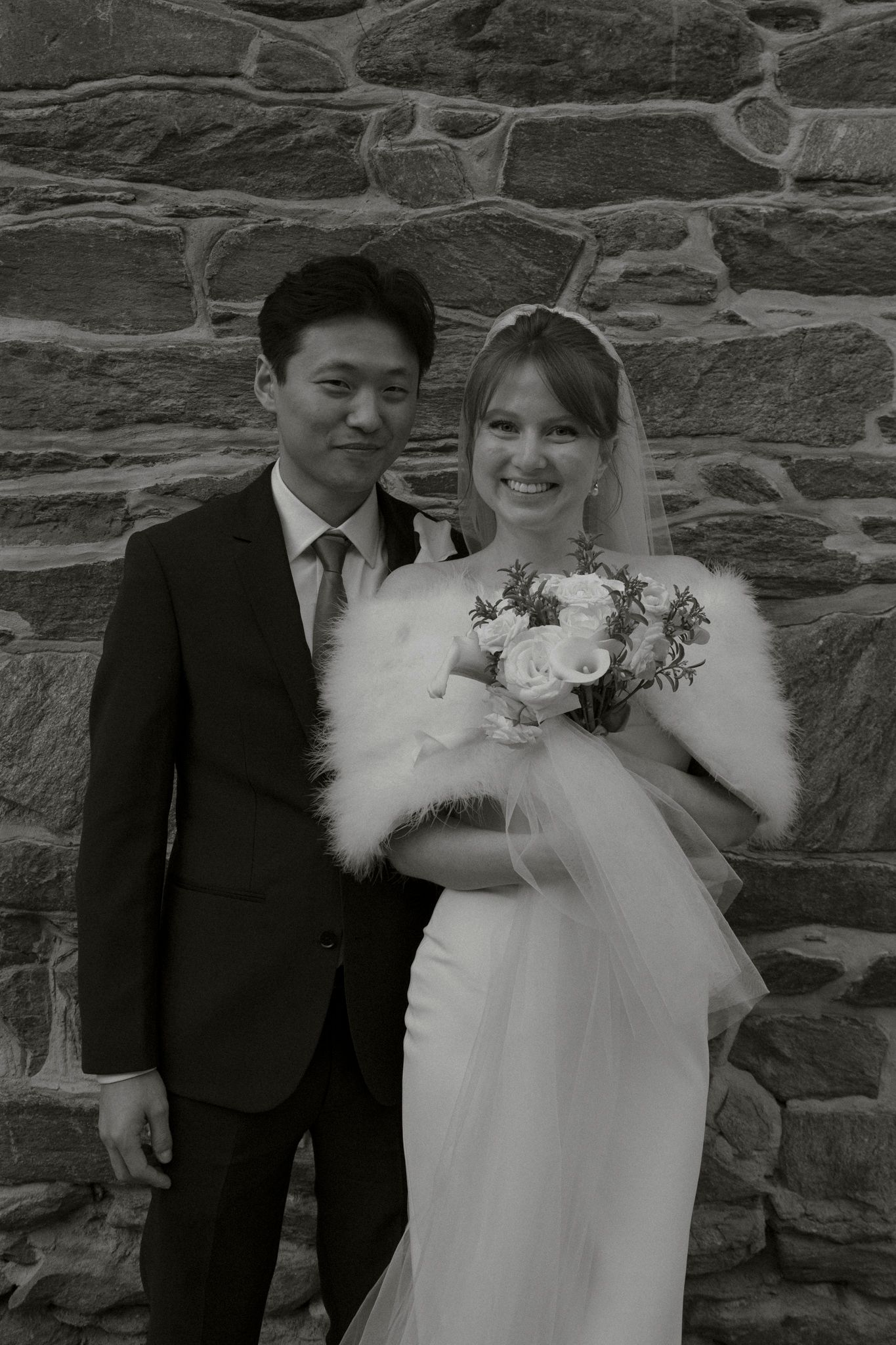 Black and white portrait of bride and groom posing before a stone wall, bride holding bouquet.