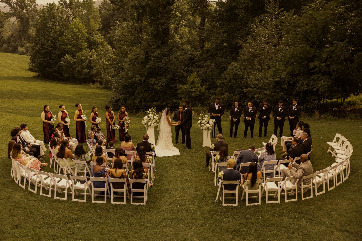 Outdoor wedding ceremony on lush green lawn with guests seated in semicircle around couple at floral arch, surrounded by trees.
