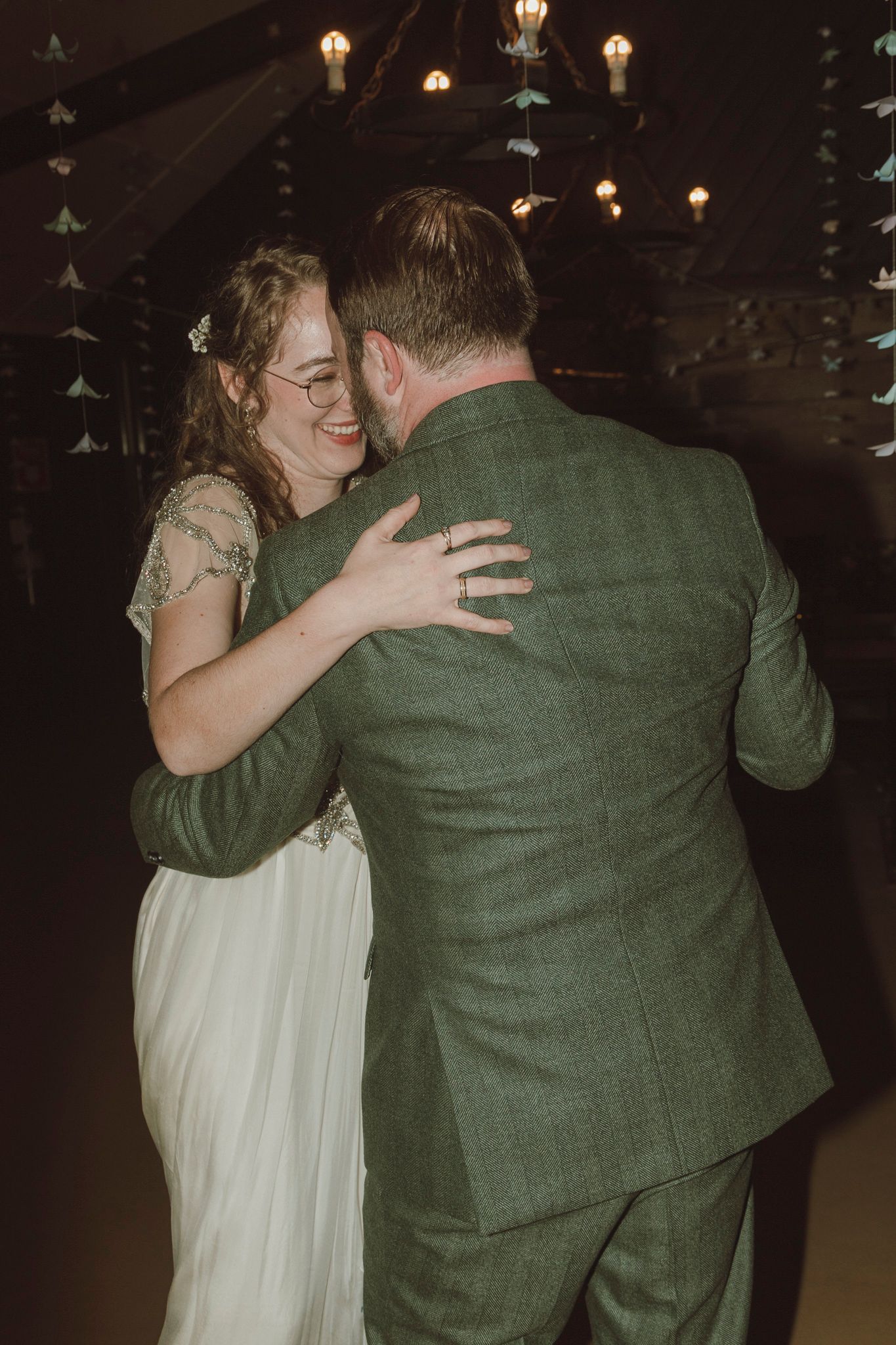 A couple shares an intimate dance moment in a dimly lit venue, with the bride in white and groom in green attire.