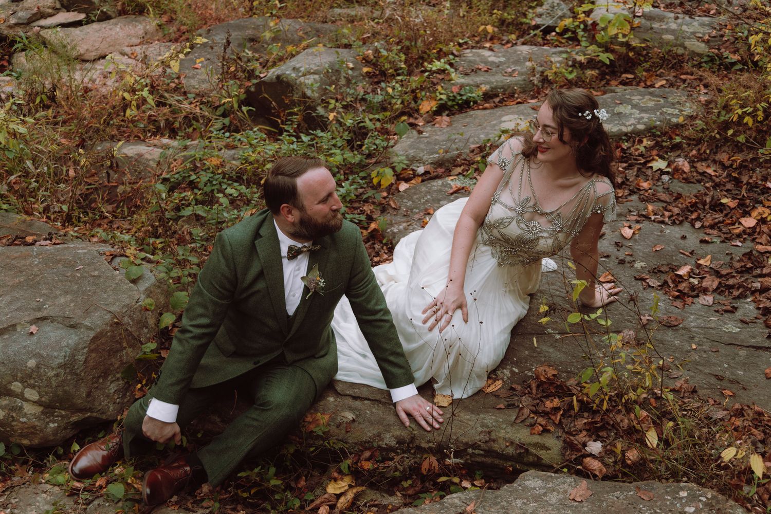 A couple in wedding attire sits on rocky terrain surrounded by autumn foliage during an outdoor photo session.