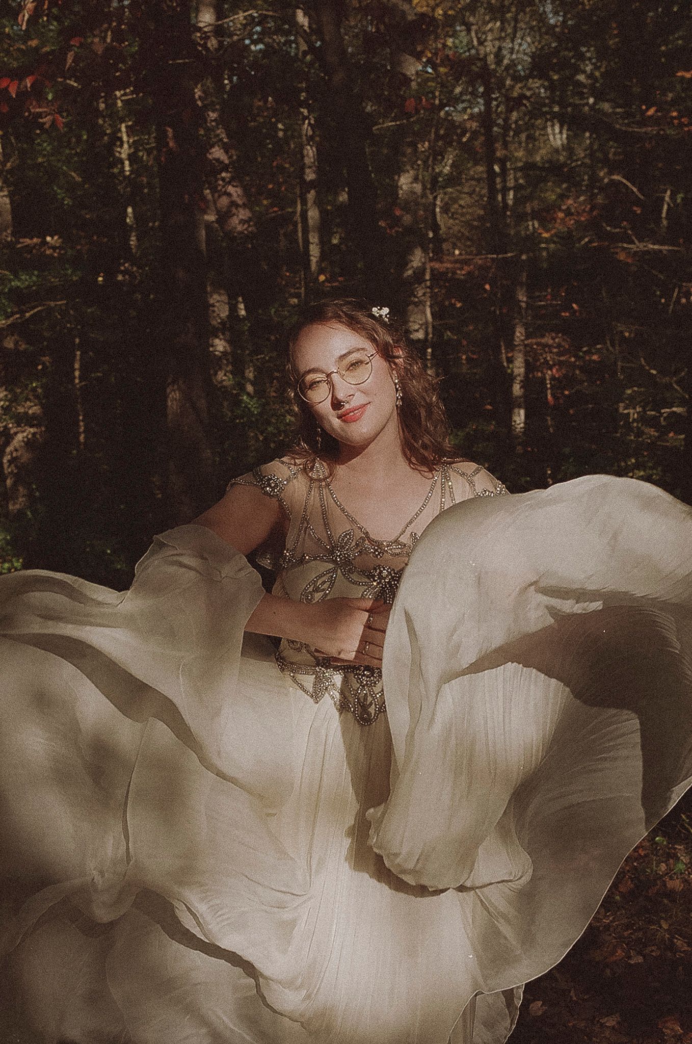 A person in a flowing white dress poses ethereally in a moody forest setting, creating a dreamy artistic portrait.