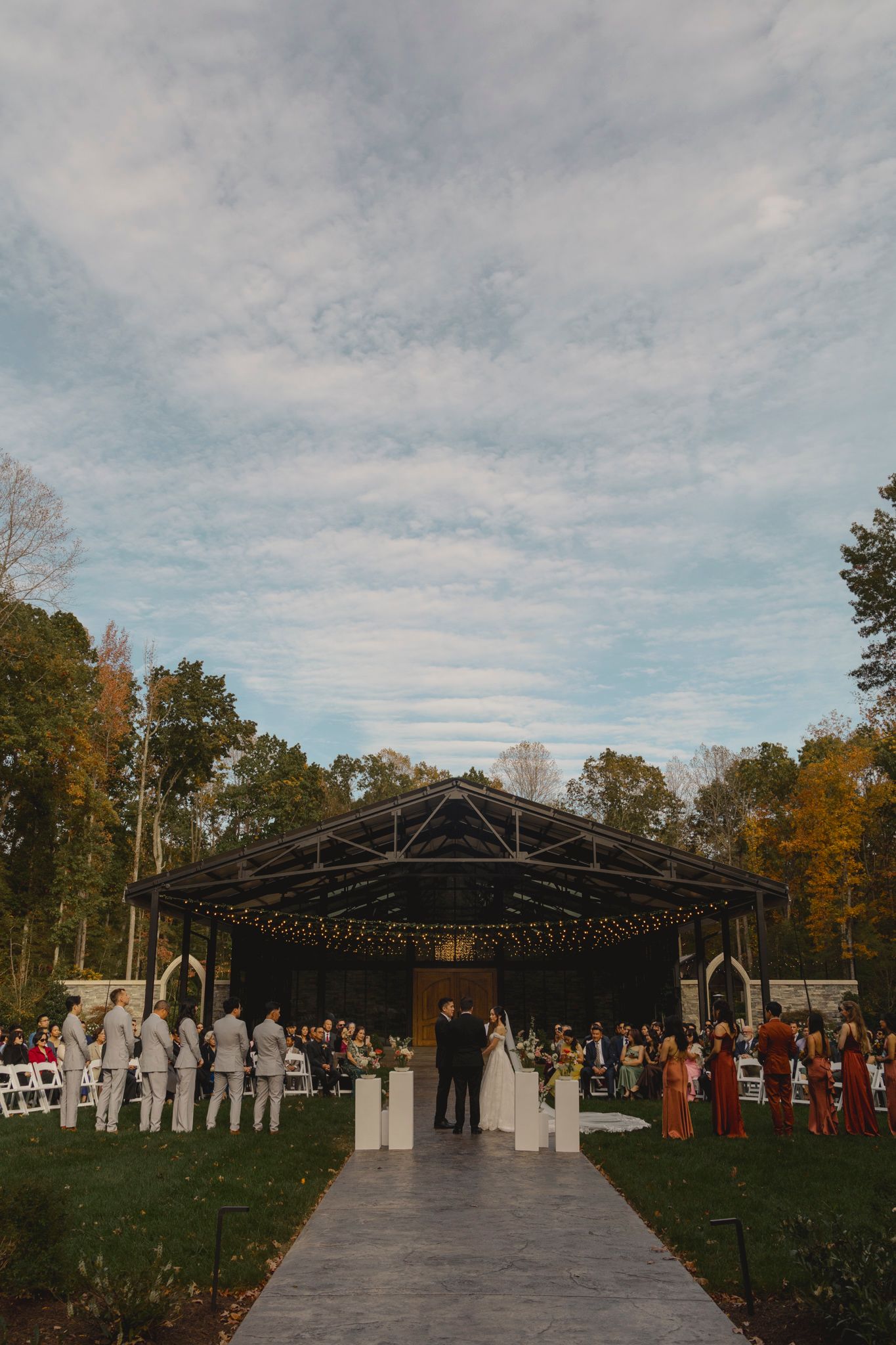 An outdoor wedding ceremony setup with a modern black pavilion structure surrounded by autumn trees and gathered guests.