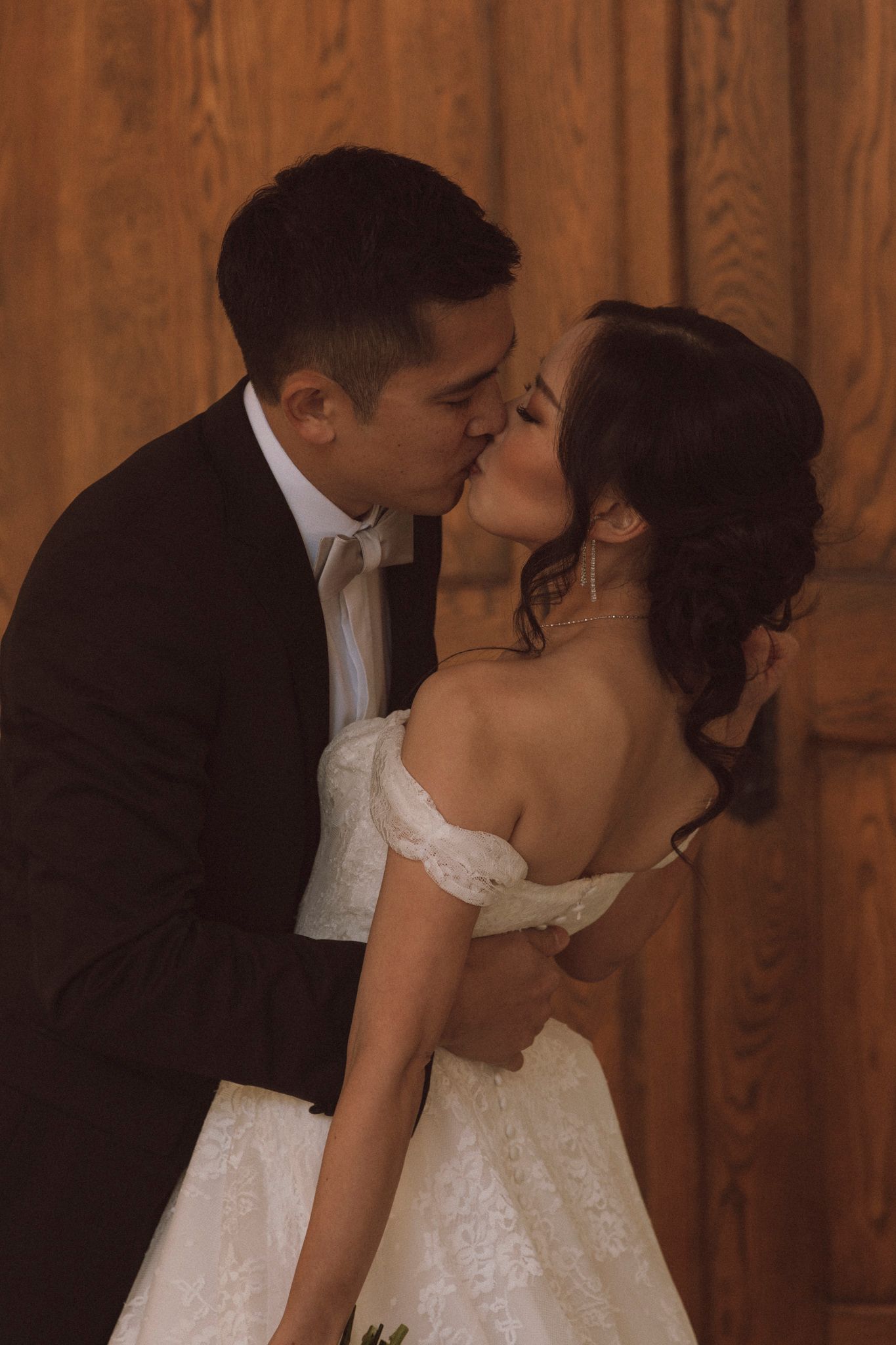 A romantic wedding moment with a couple sharing a kiss in front of a wooden backdrop.