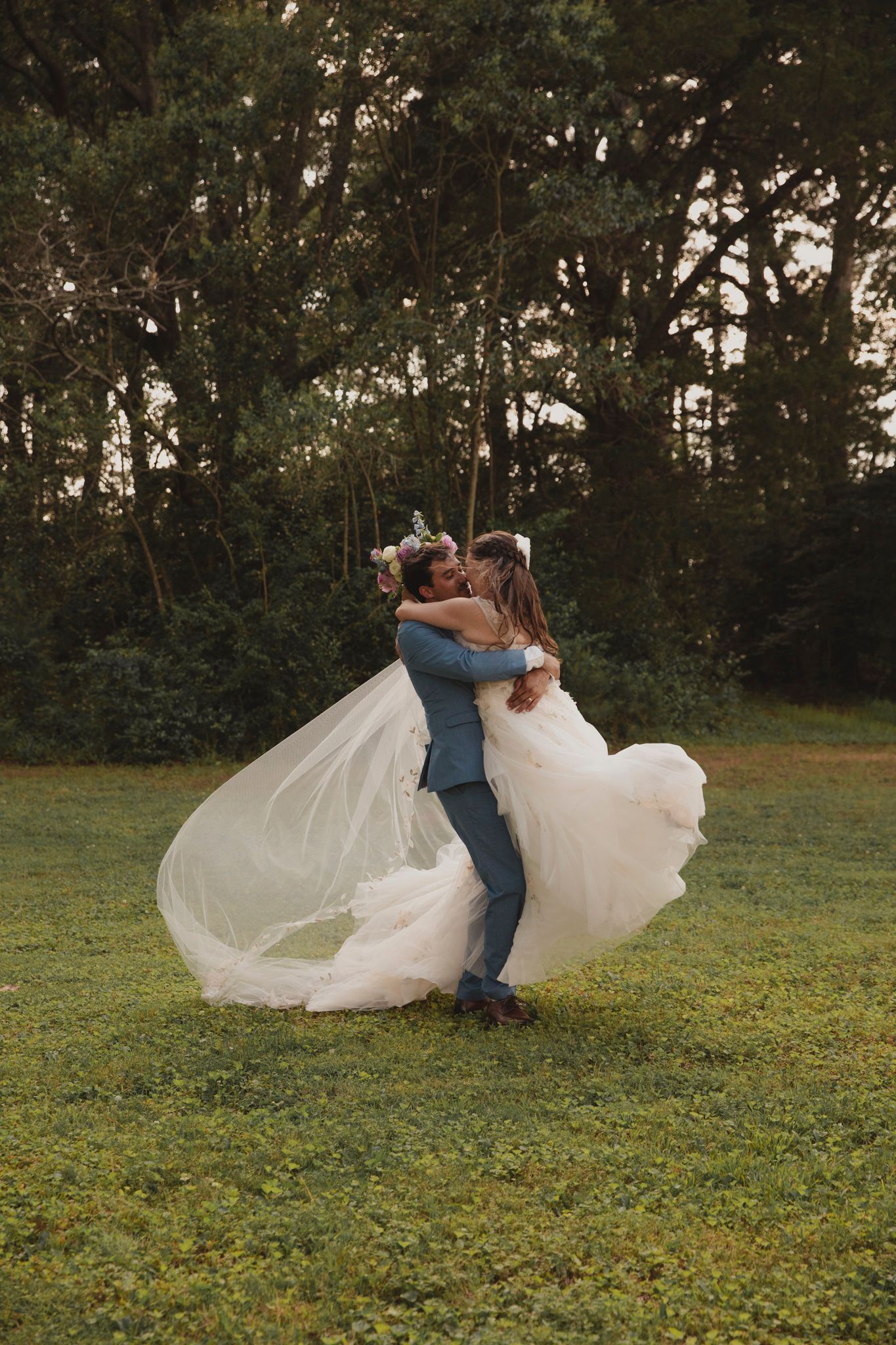A couple shares a joyful embrace on their wedding day as the white dress flows in the breeze against a wooded backdrop.