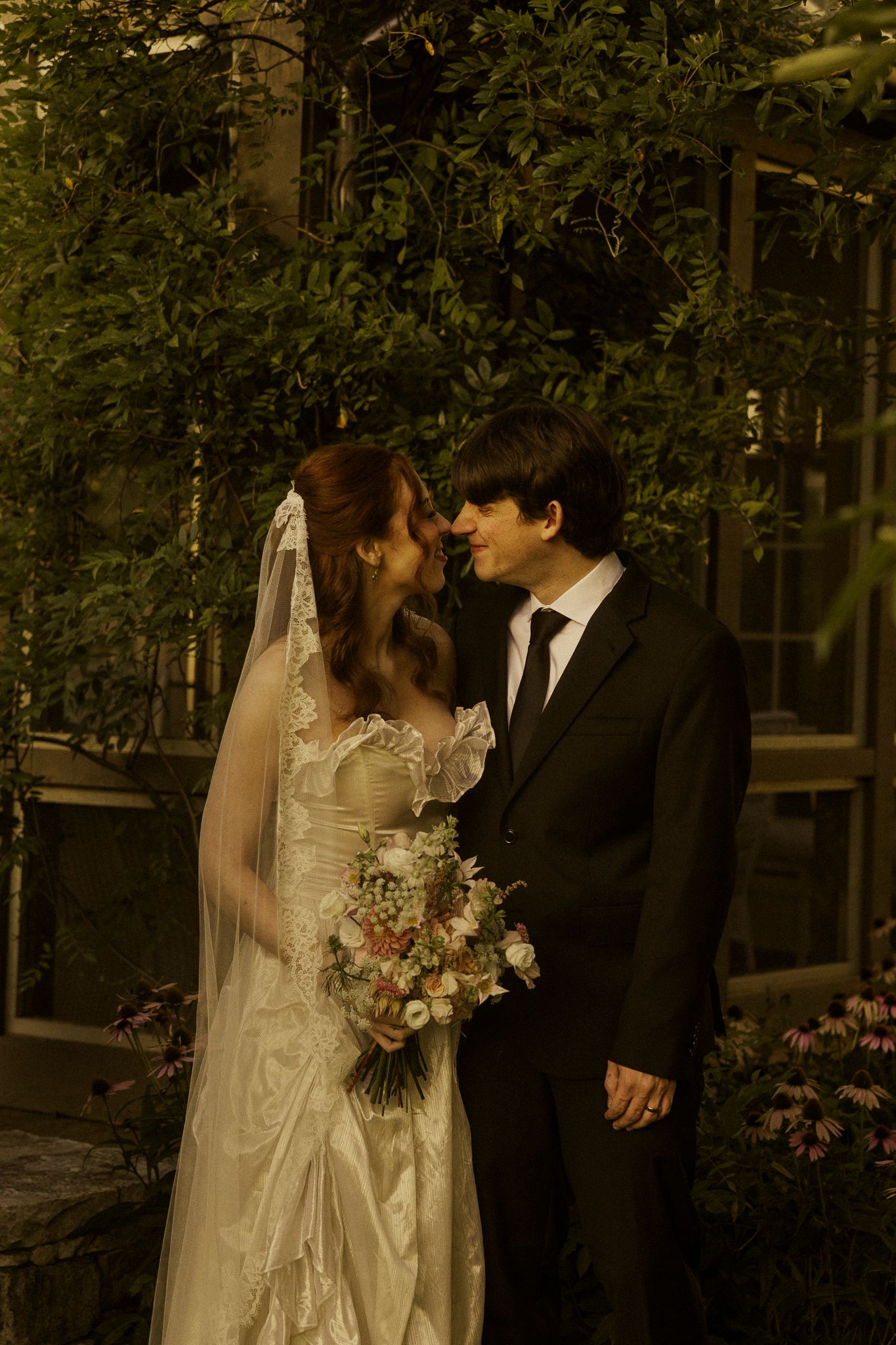 Bride and groom share a romantic moment in a lush garden, bride holding a bouquet in a satin gown with veil.