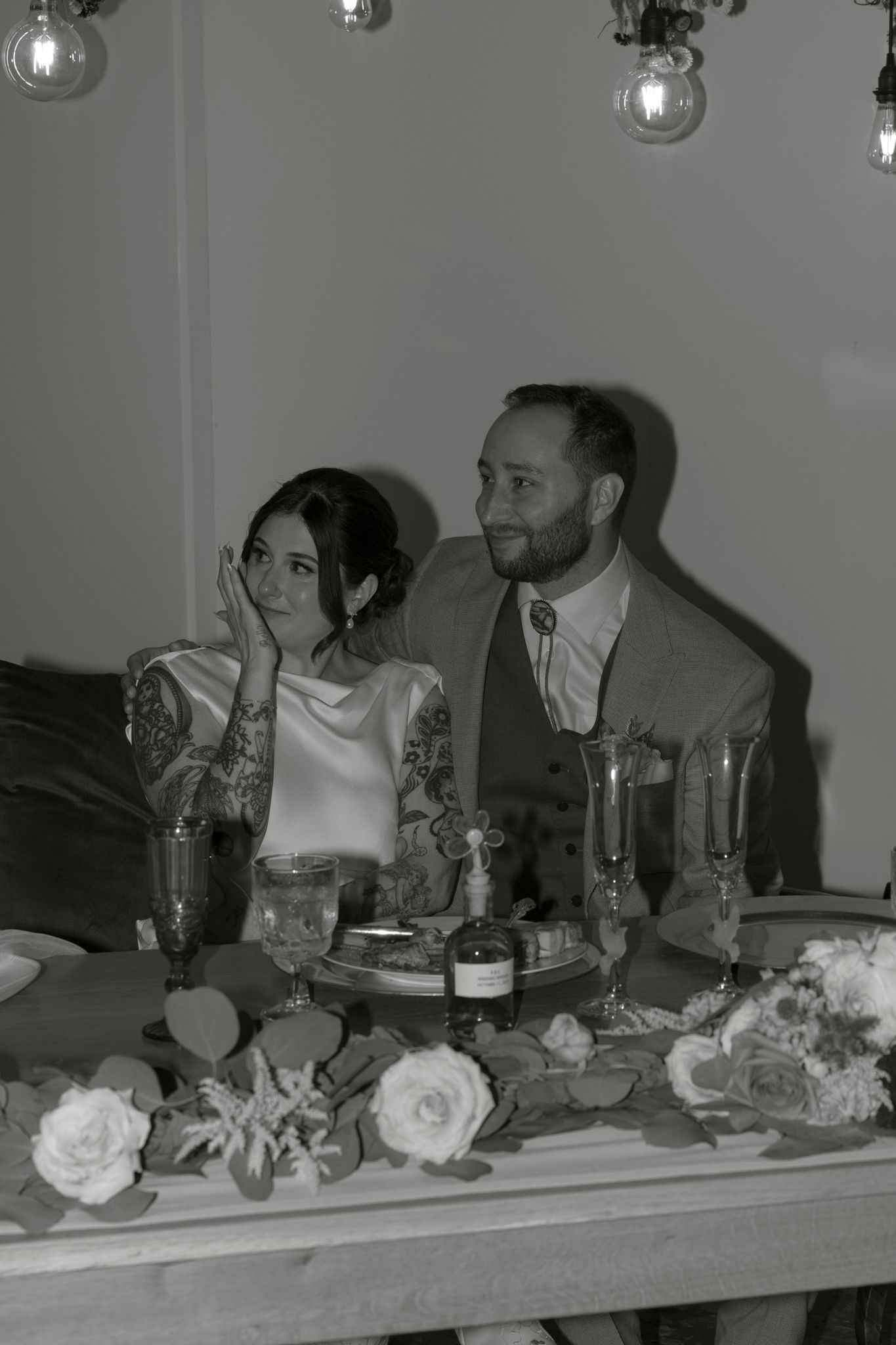 A black and white wedding reception photo showing a couple seated at a decorated table with roses and wine glasses.