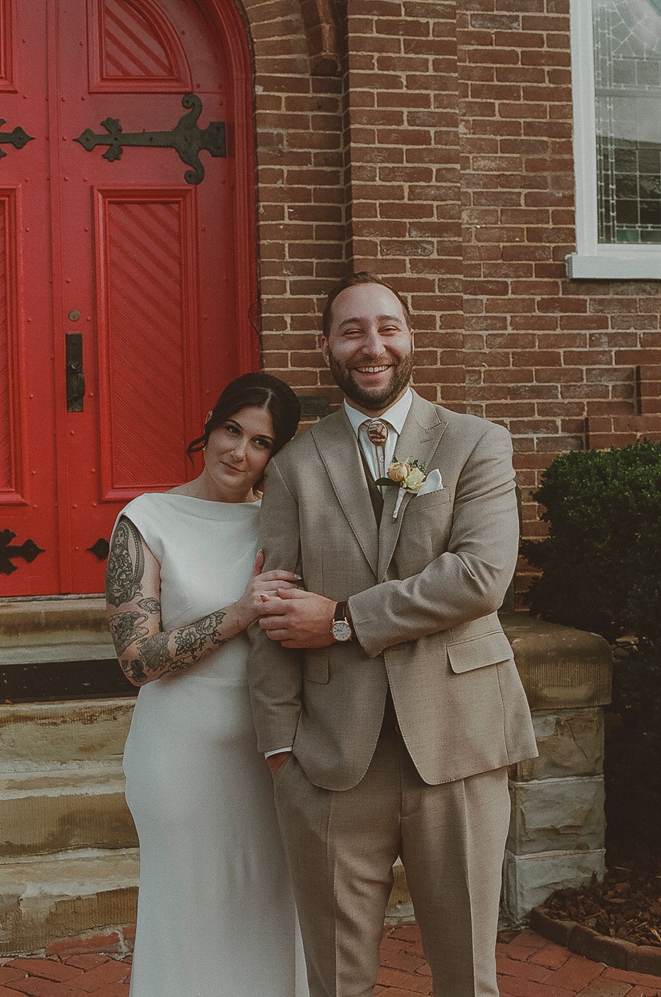 A newlywed couple shares a joyful moment in front of a brick building with bright red doors.