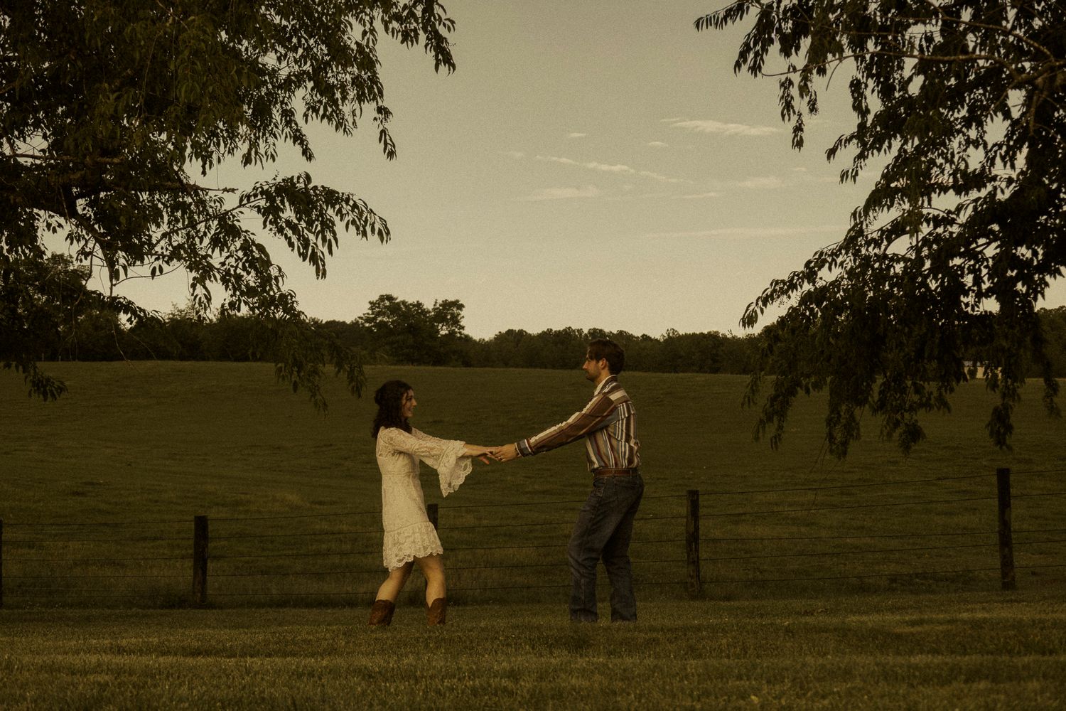 Couple dancing hand in hand in a lush green field at dusk under tree branches.