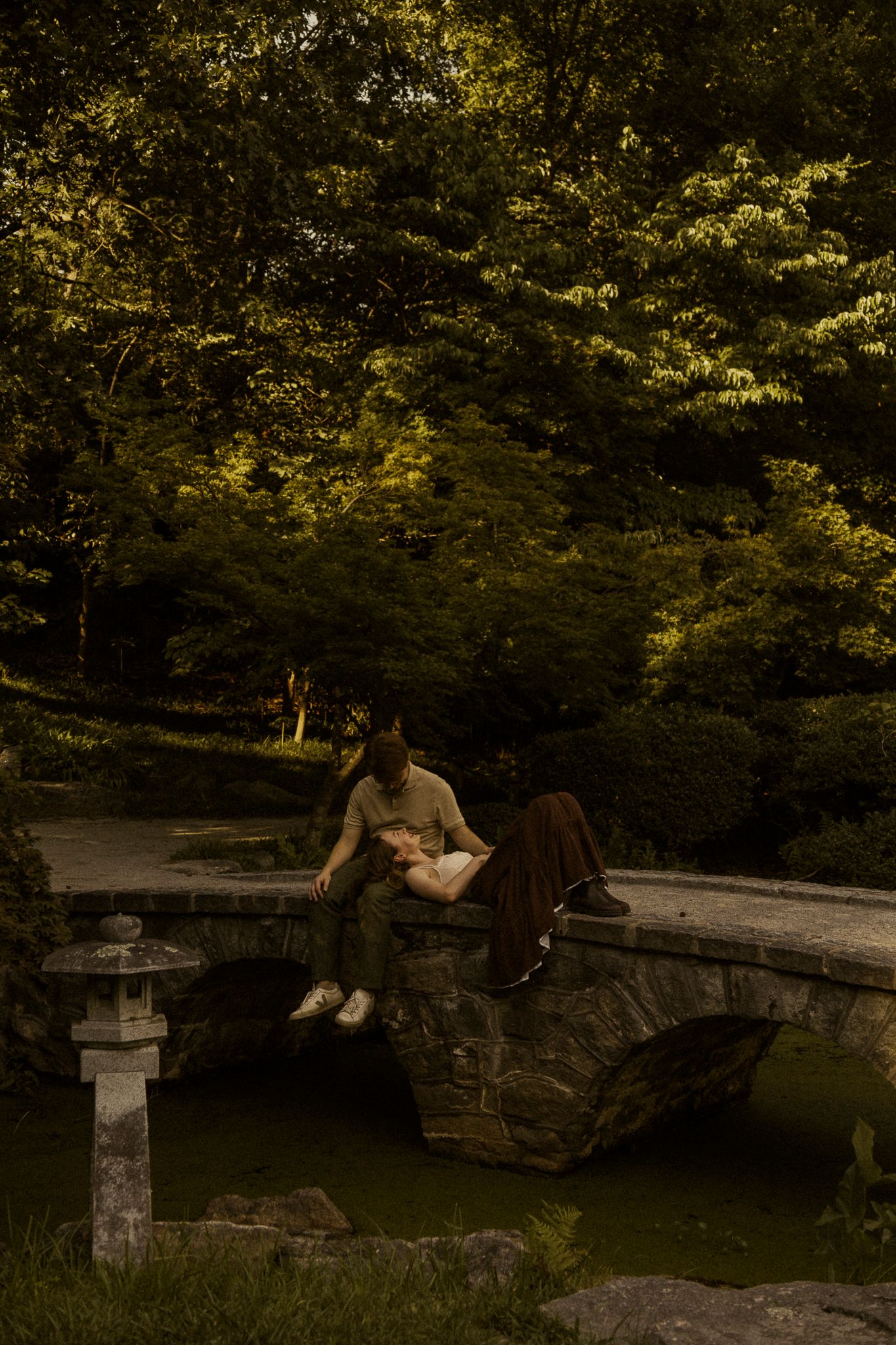 Couple relaxing on a stone bridge in a lush Japanese garden, surrounded by green trees near a tranquil pond.