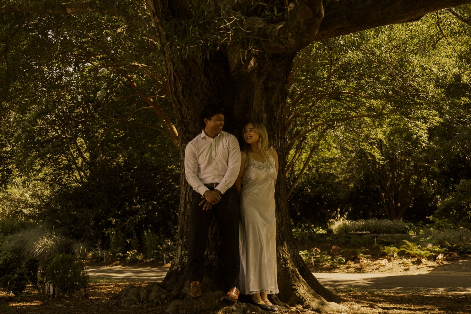 Couple standing together under a large tree in a sunlit garden, woman in white dress leaning on man.