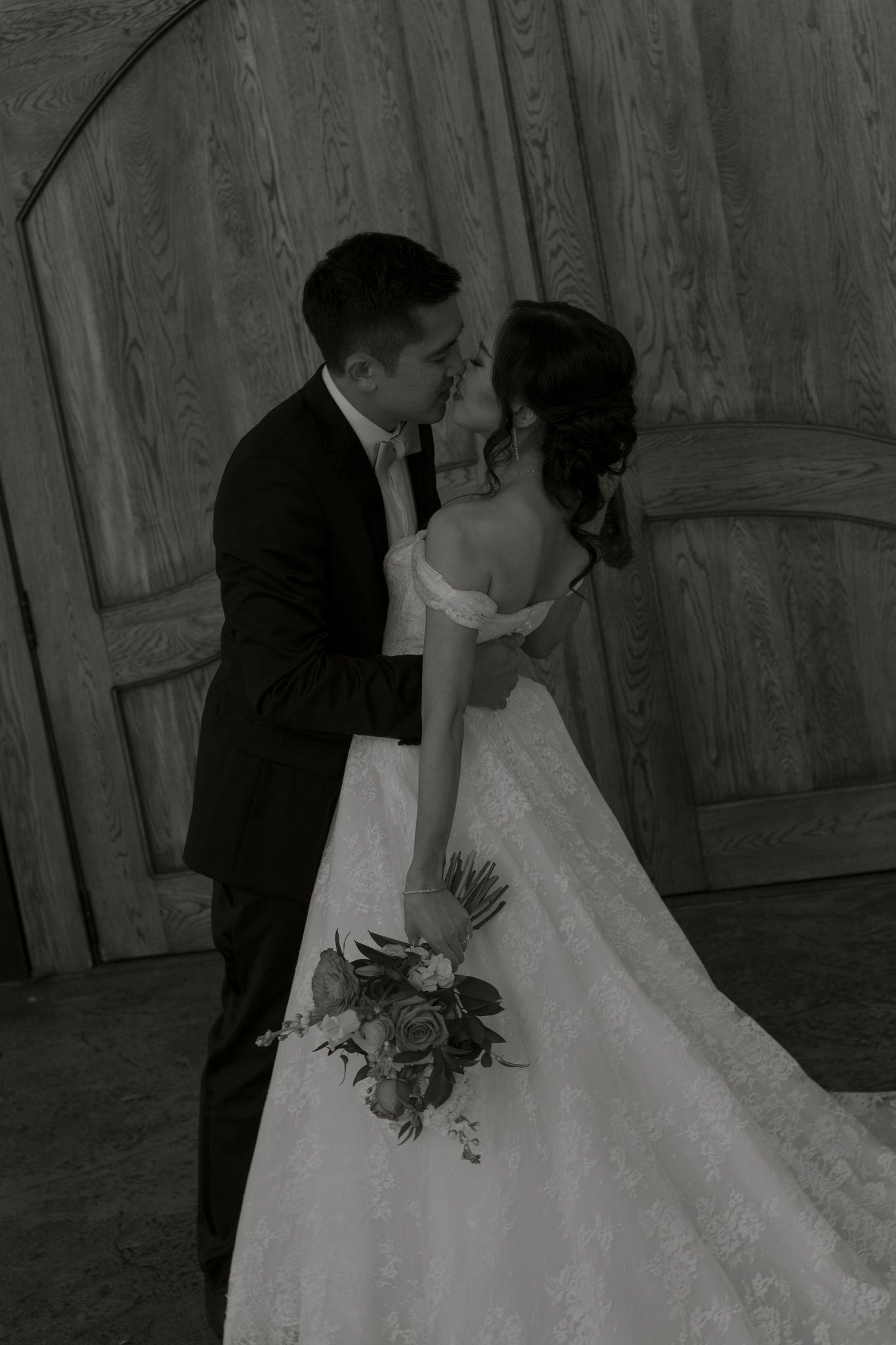 A romantic black and white wedding photo series showing a couple kissing in front of rustic wooden barn doors.