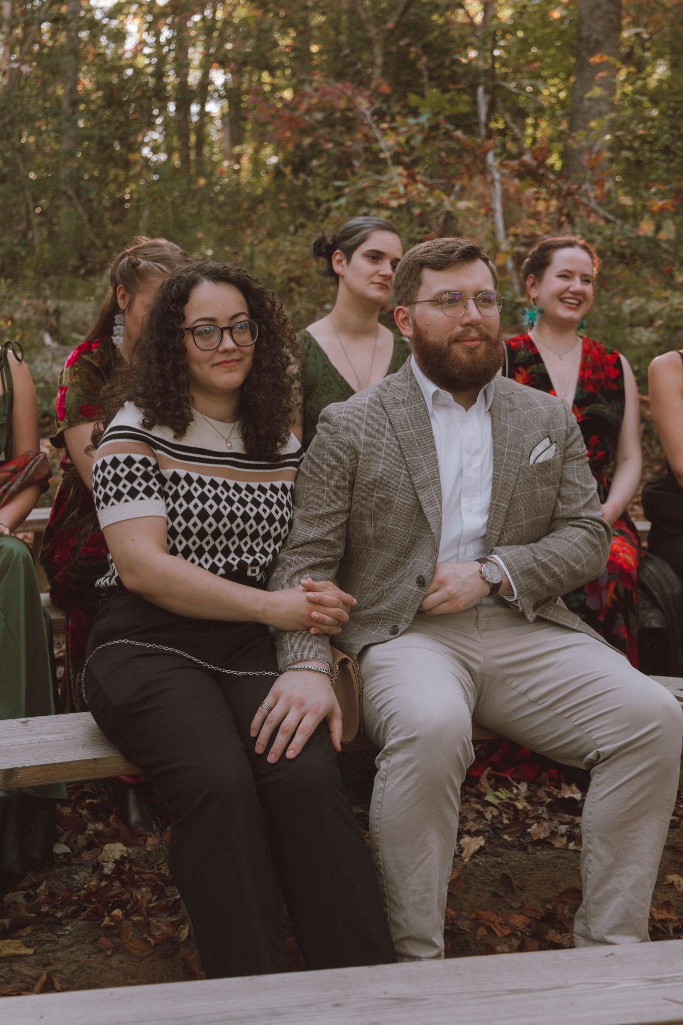 Group of friends sit casually on wooden benches in an autumn forest setting wearing coordinated casual attire.