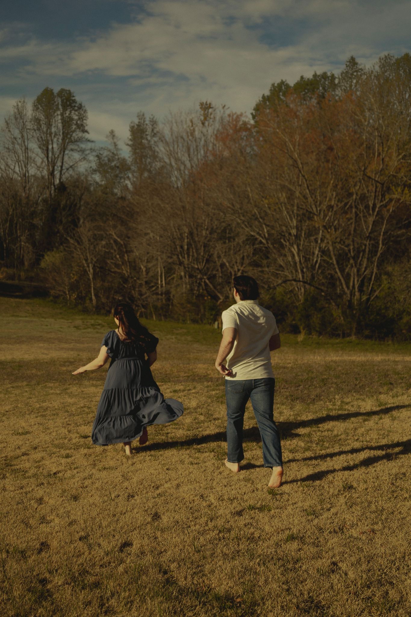 Barefoot couple running hand-in-hand across a sunlit meadow with fall trees.