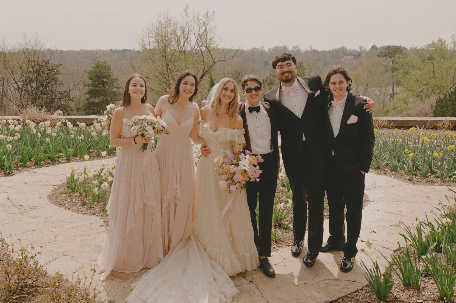 Wedding party in formal attire poses together outdoors in a natural desert landscape setting.