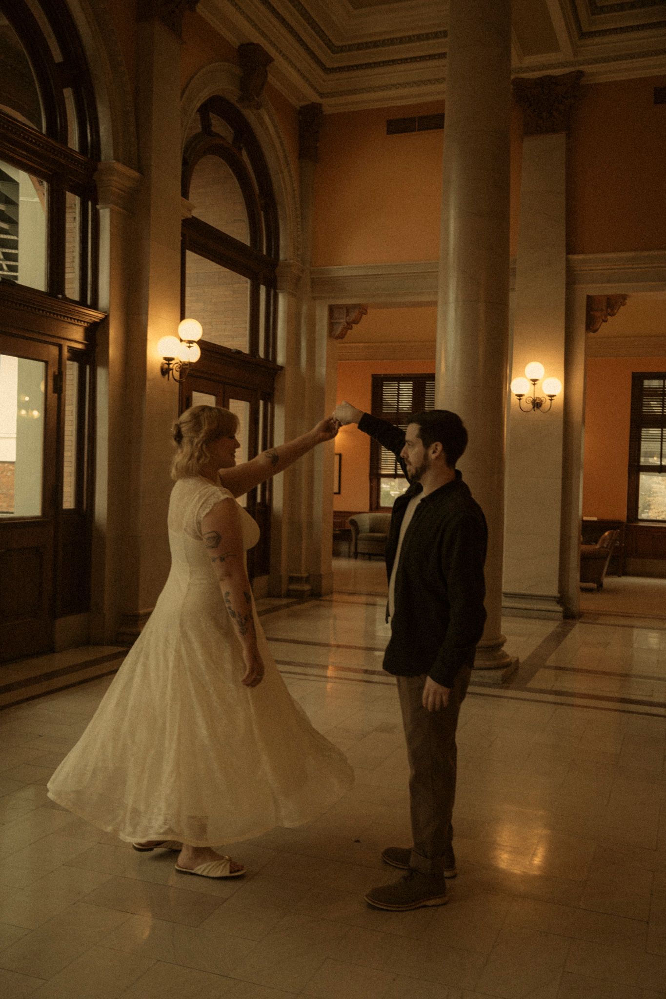 A couple shares a romantic dance in an elegant hallway with ornate columns and warm lighting.