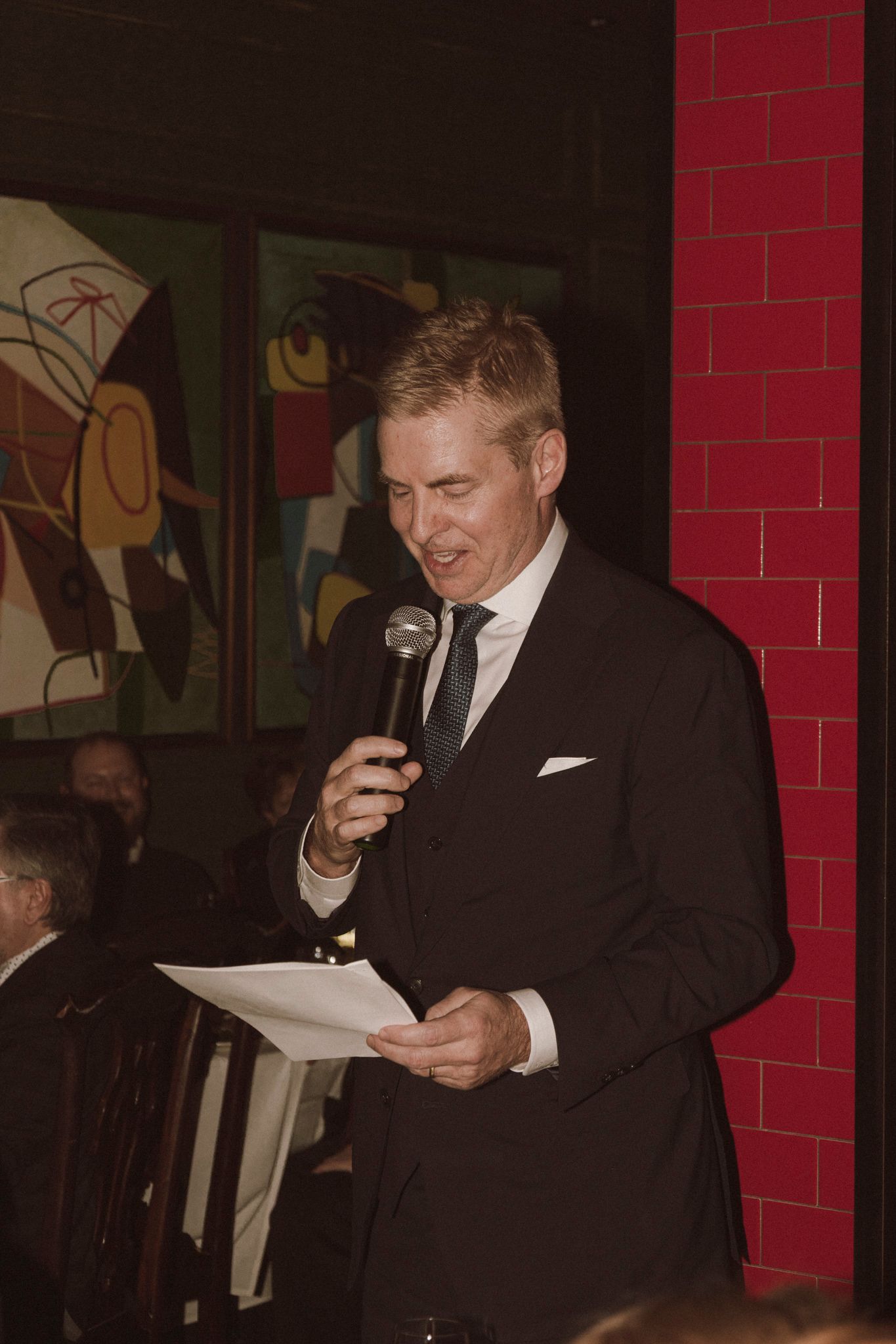 Blond man in black suit delivers a wedding toast holding microphone and paper in front of artwork.