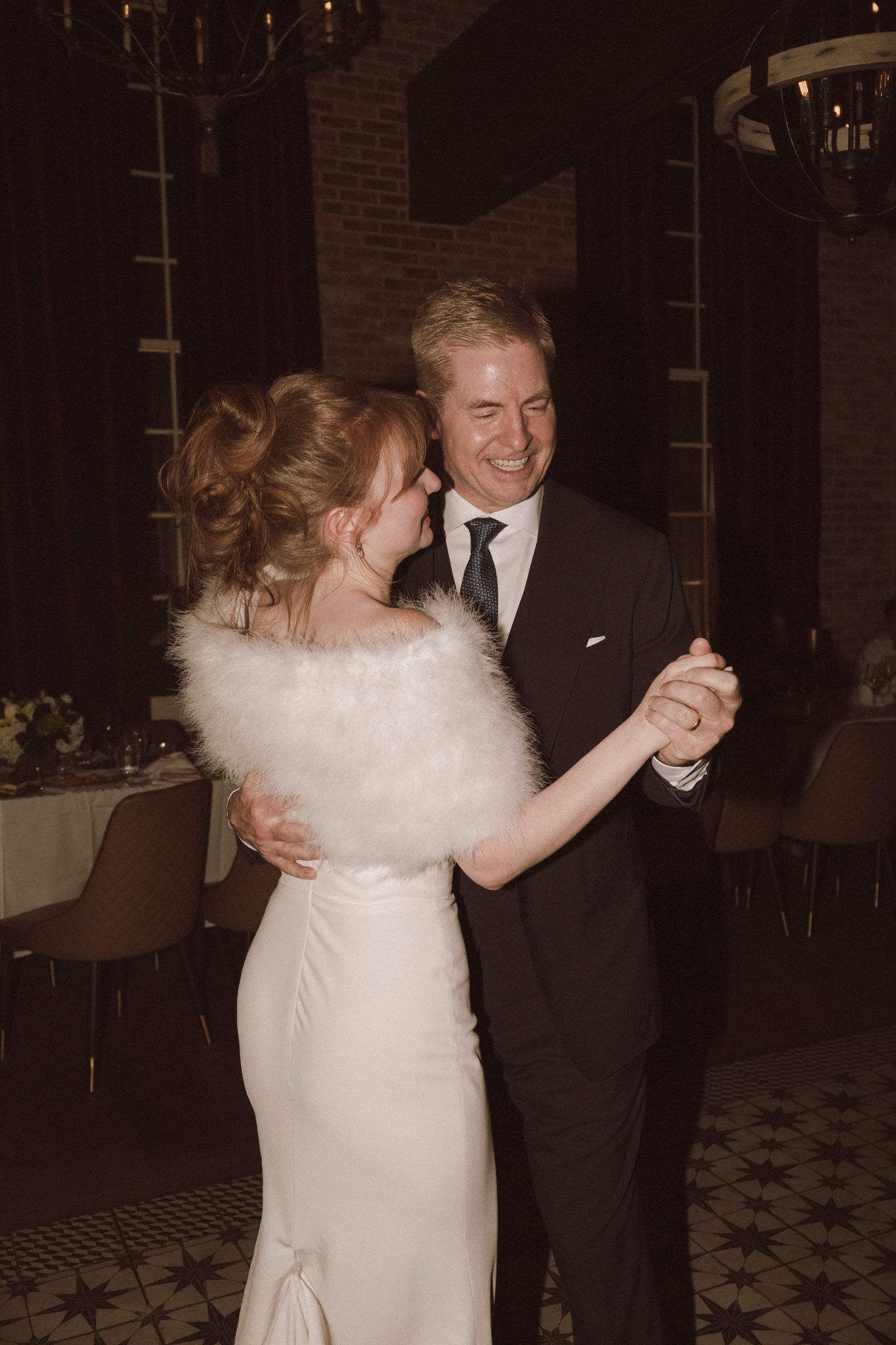 Bride and groom share their first dance, smiling joyfully in elegant wedding attire at a warmly lit reception.
