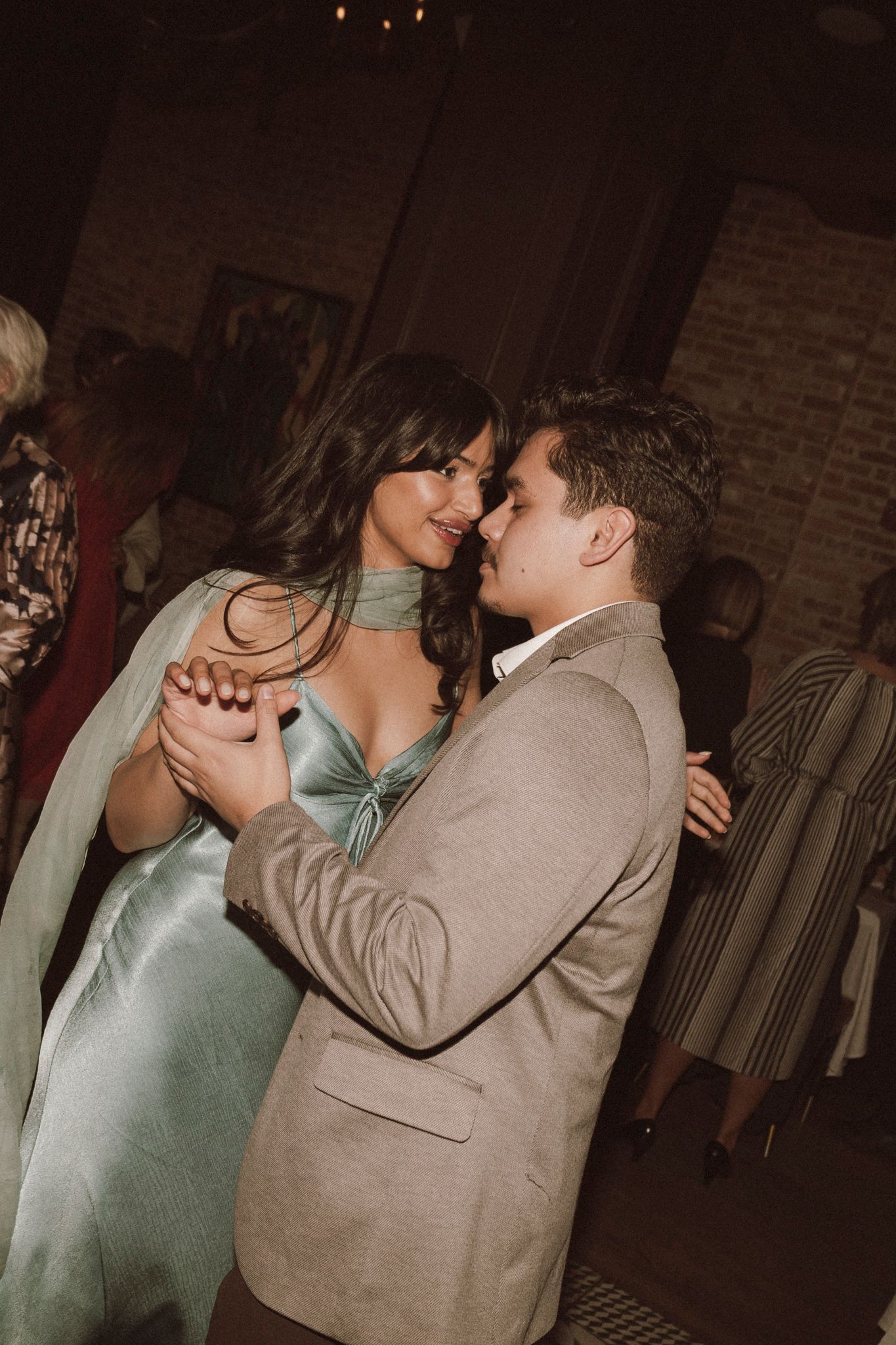 A couple shares a romantic slow dance, the woman in a sage green satin gown at an elegant reception.