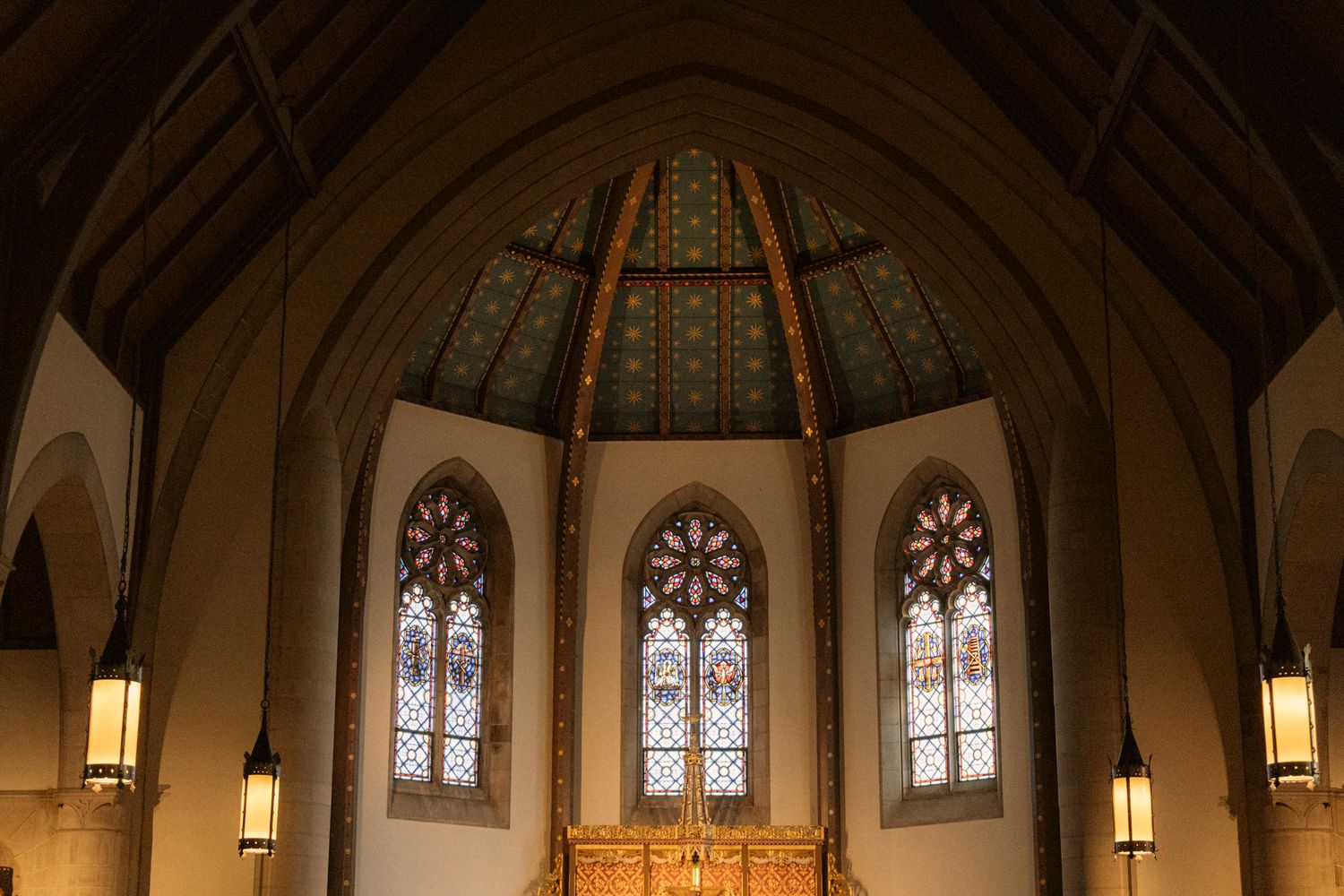 Gothic church interior with ornate stained glass windows above a golden altar beneath an octagonal vaulted ceiling.