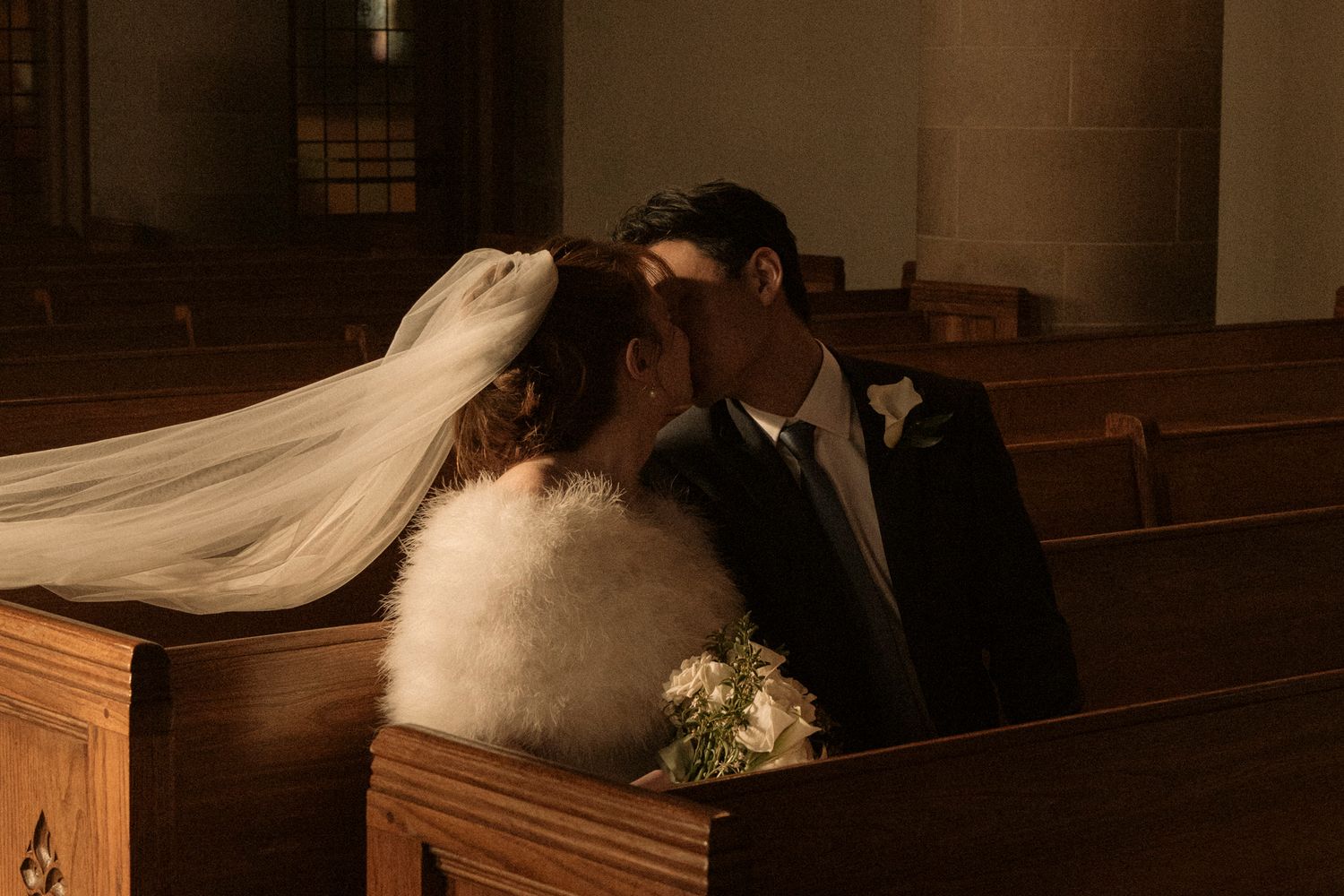 Bride and groom share a romantic kiss in a warmly lit church pew, bridal veil flowing, holding a bouquet.