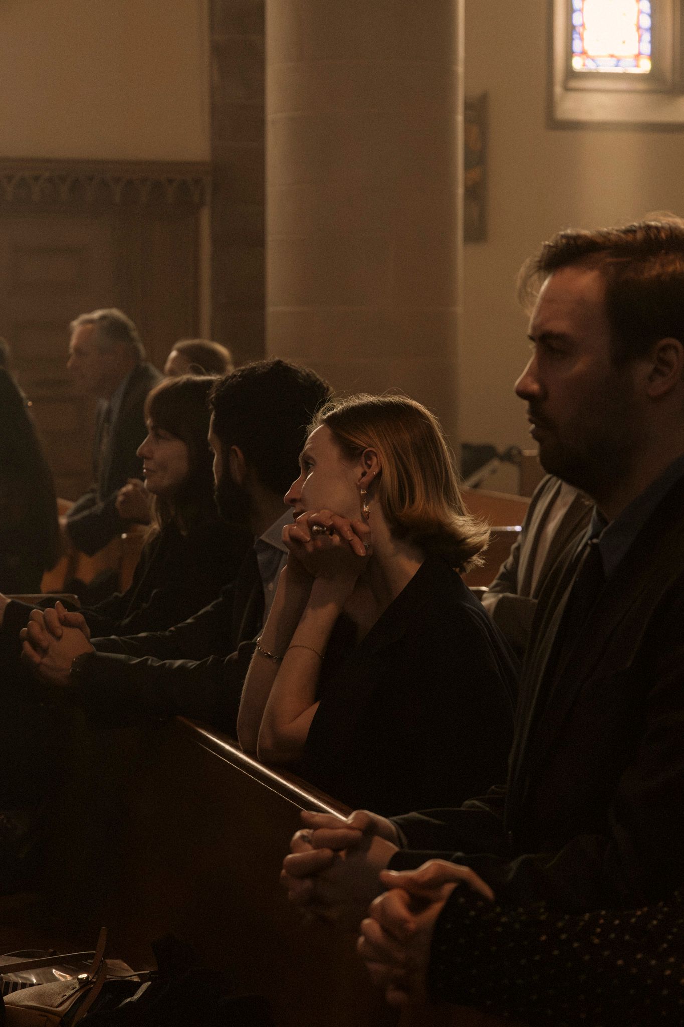 Wedding guests seated in dimly lit church pews, with a woman in black dress and man in suit attentively watching ceremony.