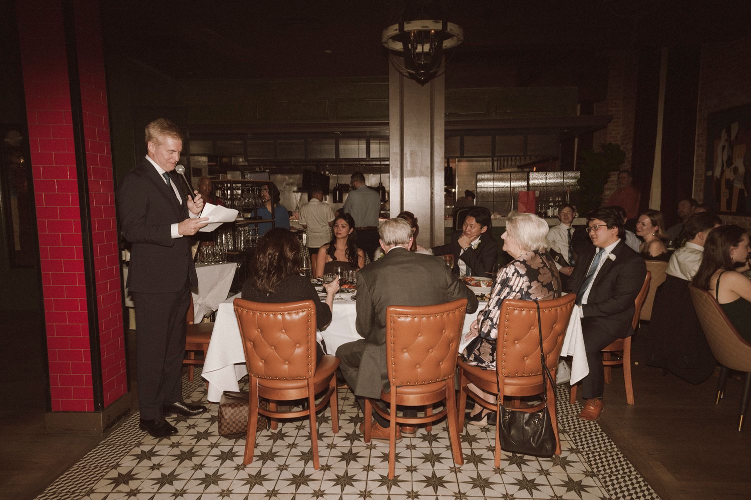 Wedding speaker addresses seated guests in an elegant restaurant with patterned tile floors.