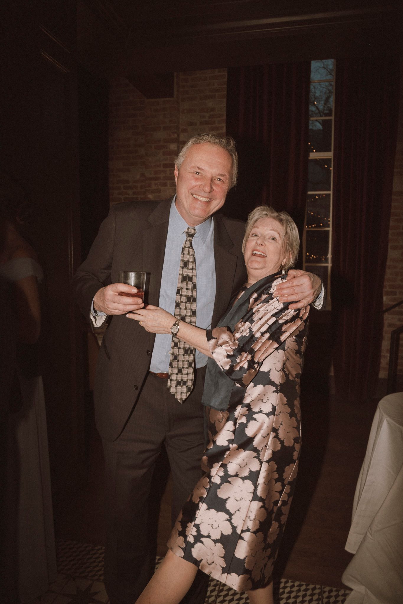Smiling couple embracing at elegant wedding reception, man in suit holding drink, woman in floral dress.