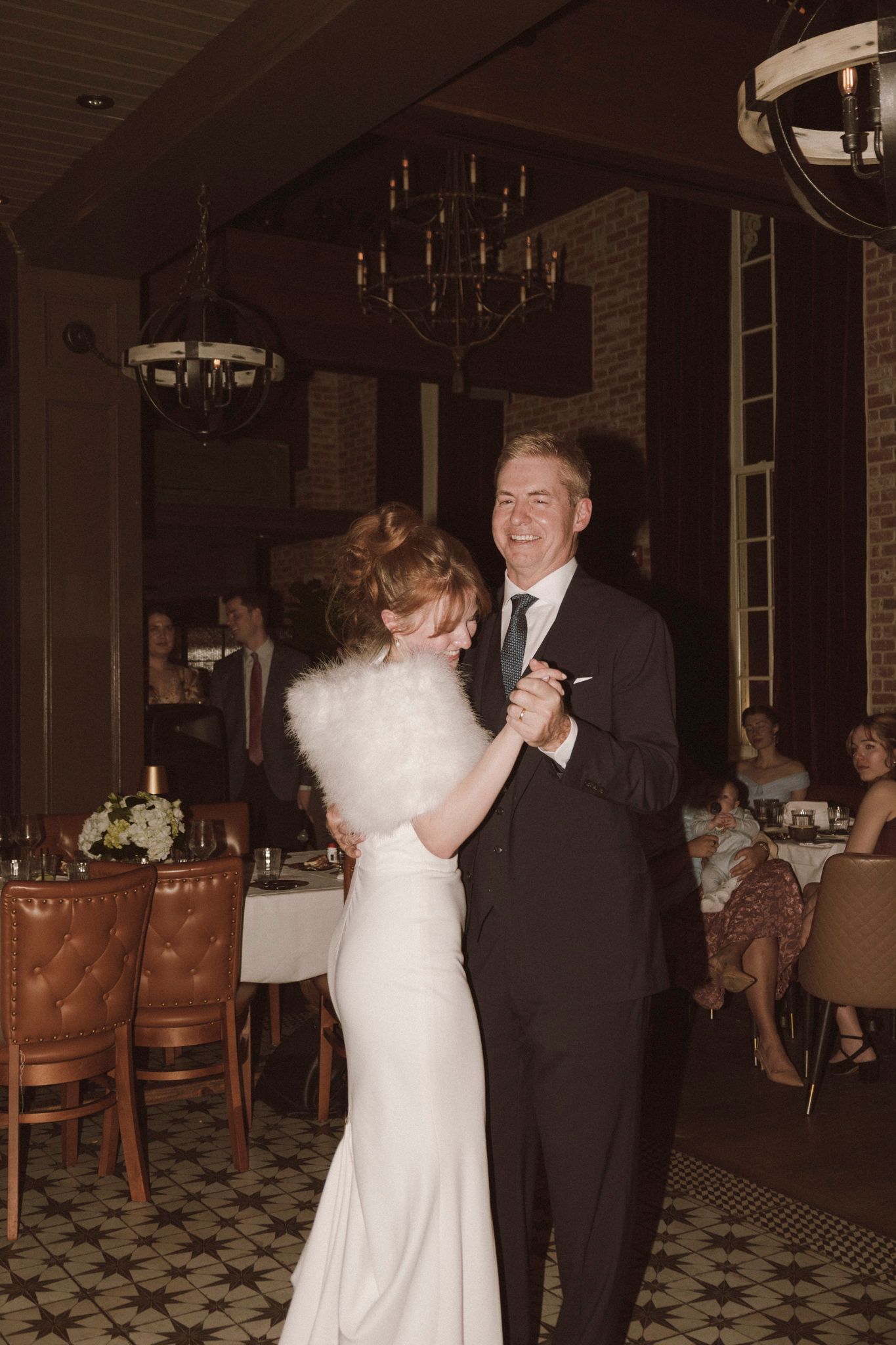 Bride and groom share first dance in elegant restaurant venue with chandelier and leather chairs.