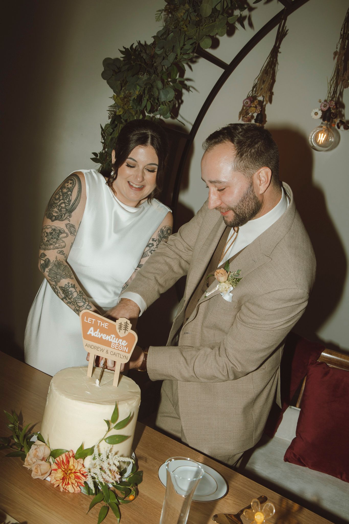 A couple cuts into their elegant white wedding cake decorated with fresh flowers at their reception.