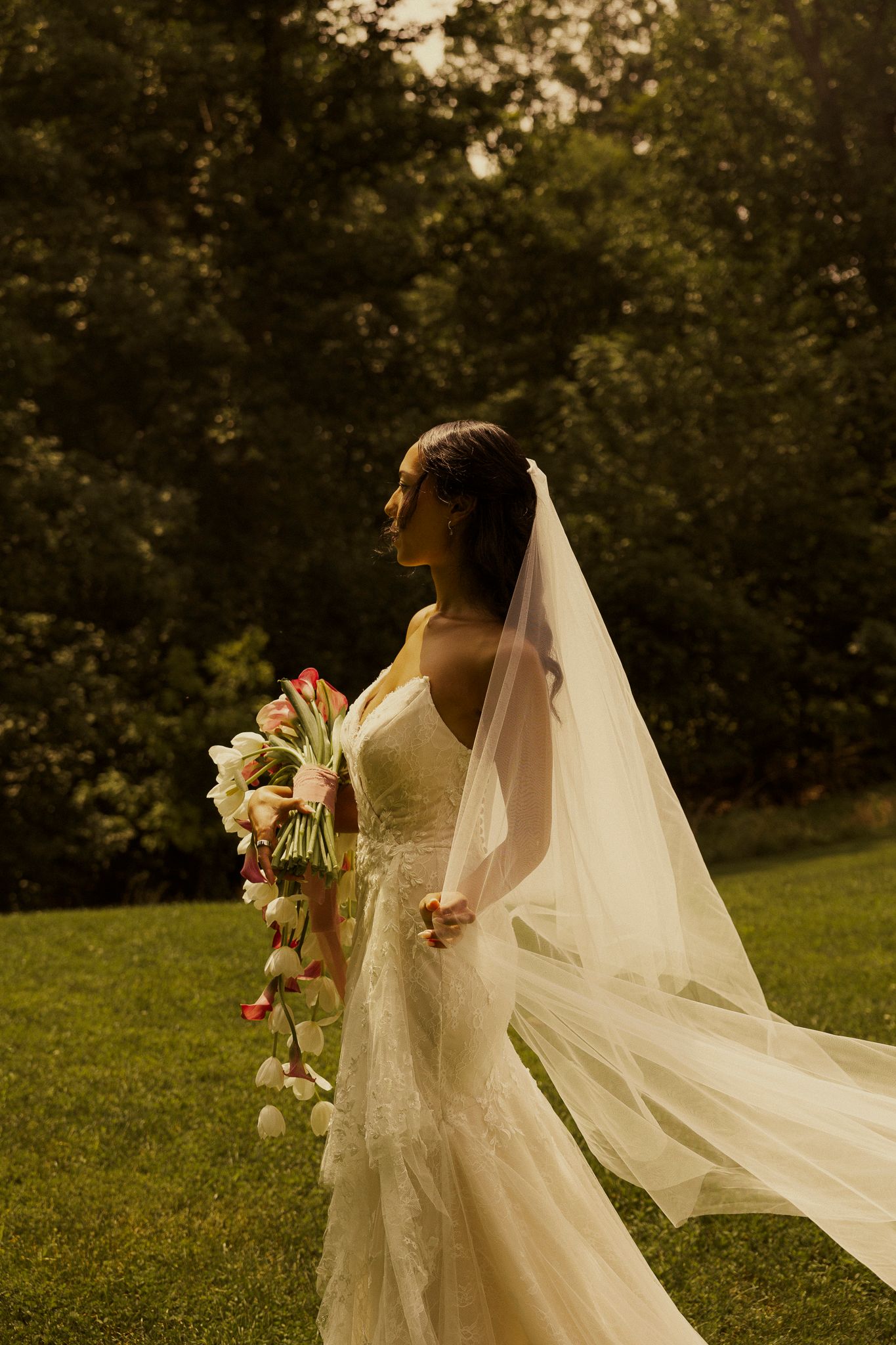 Bride in lace mermaid gown with flowing veil holds floral bouquet outdoors in golden light.