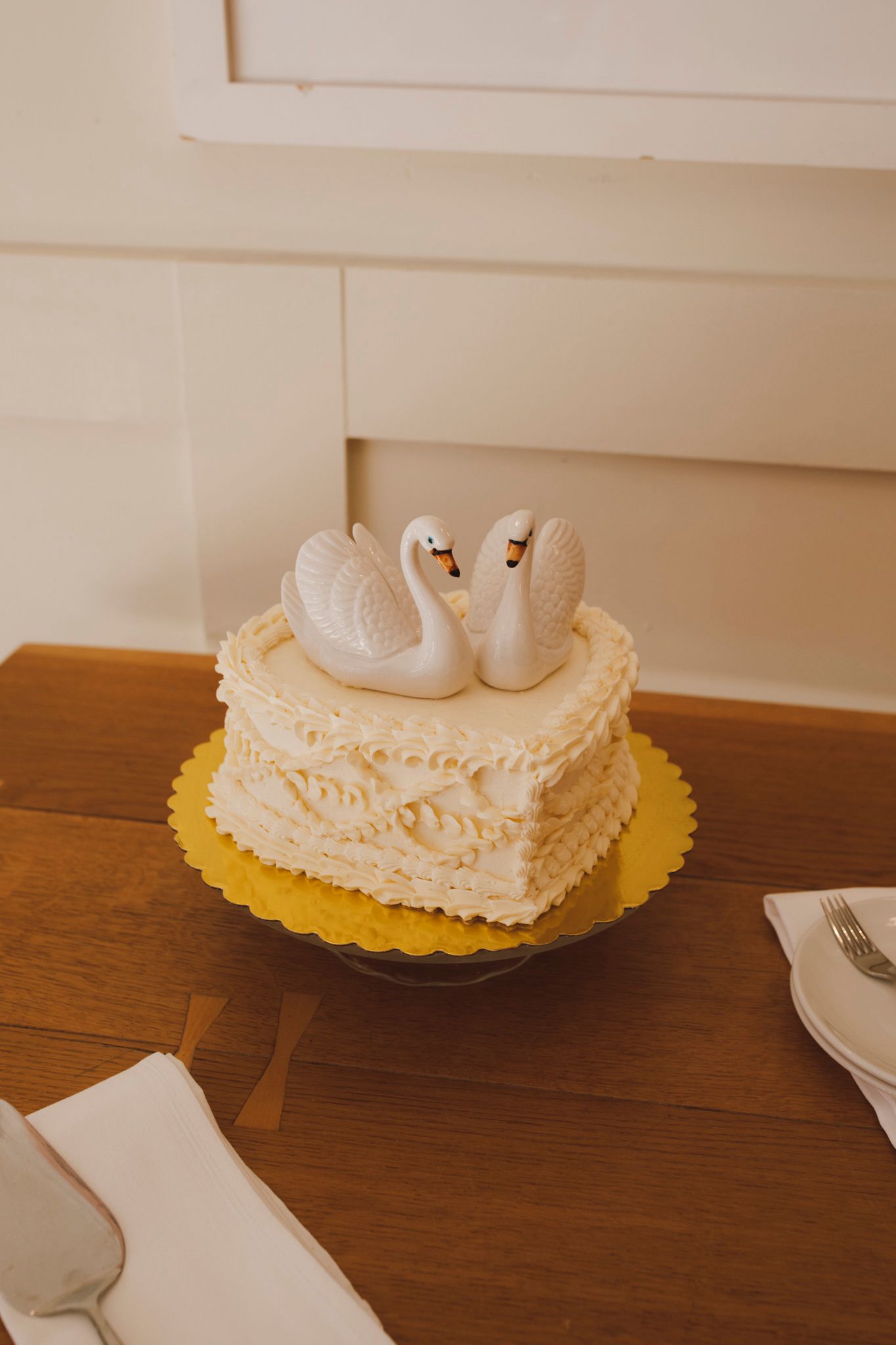 A white frosted wedding cake decorated with delicate swan figures sits on a yellow cake board on a wooden table.
