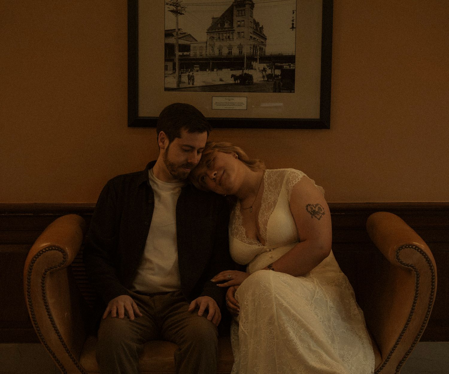 A couple in wedding attire sits close together on a vintage brown couch in a dimly lit room with framed artwork.