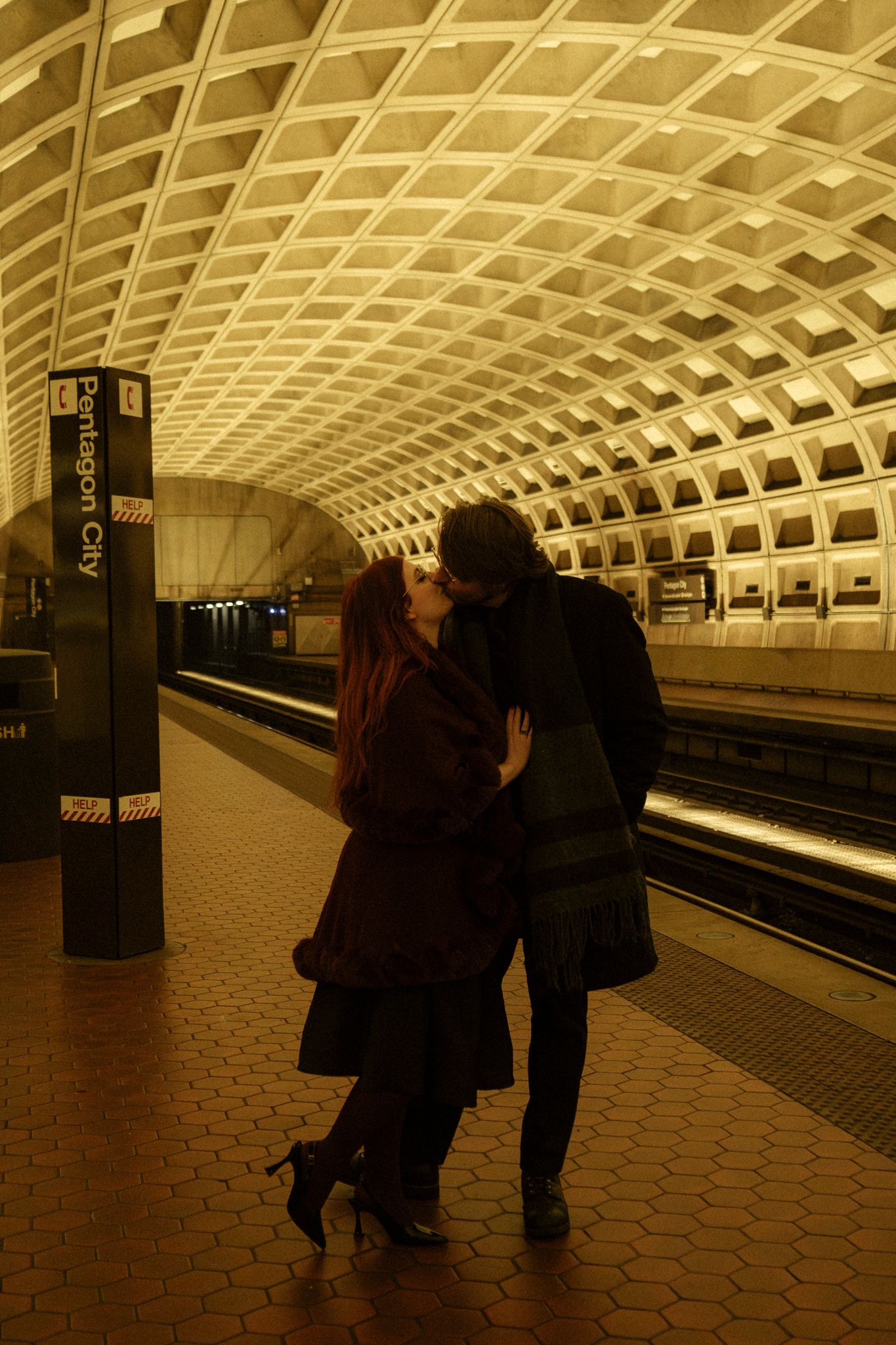 Silhouettes of people waiting on a subway platform with distinctive vaulted ceiling and hexagonal tile floor in sepia tones.