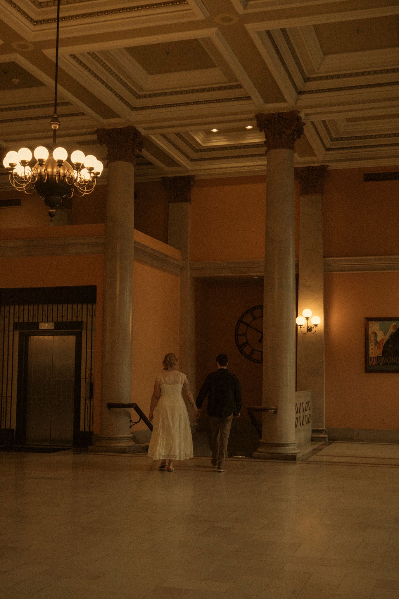 Elegant ballroom interior with ornate ceiling, chandeliers and a wedding dress display in warm ambient lighting.