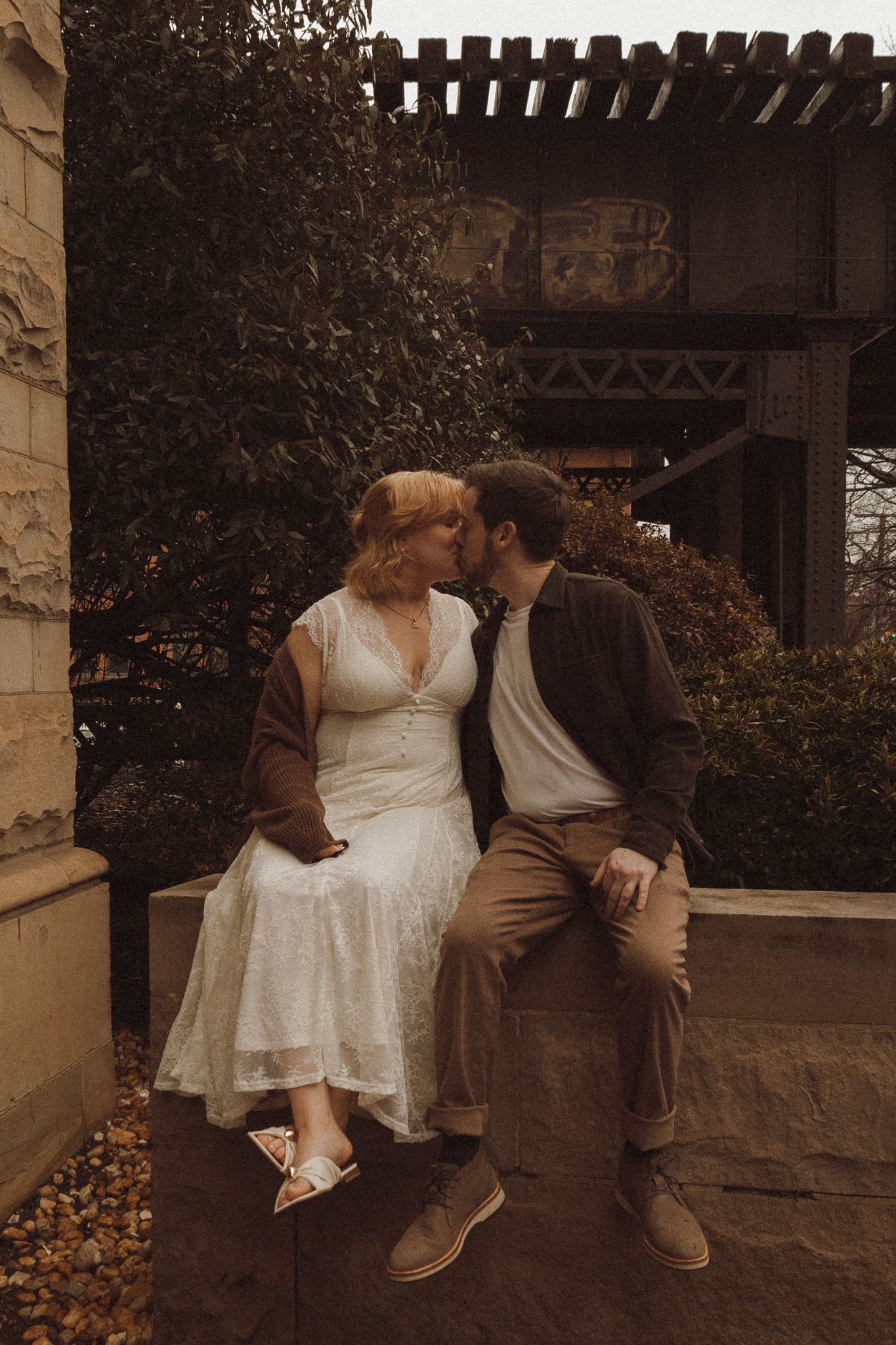 A couple in vintage-style clothing share an intimate moment on a stone wall beneath an autumn pergola.