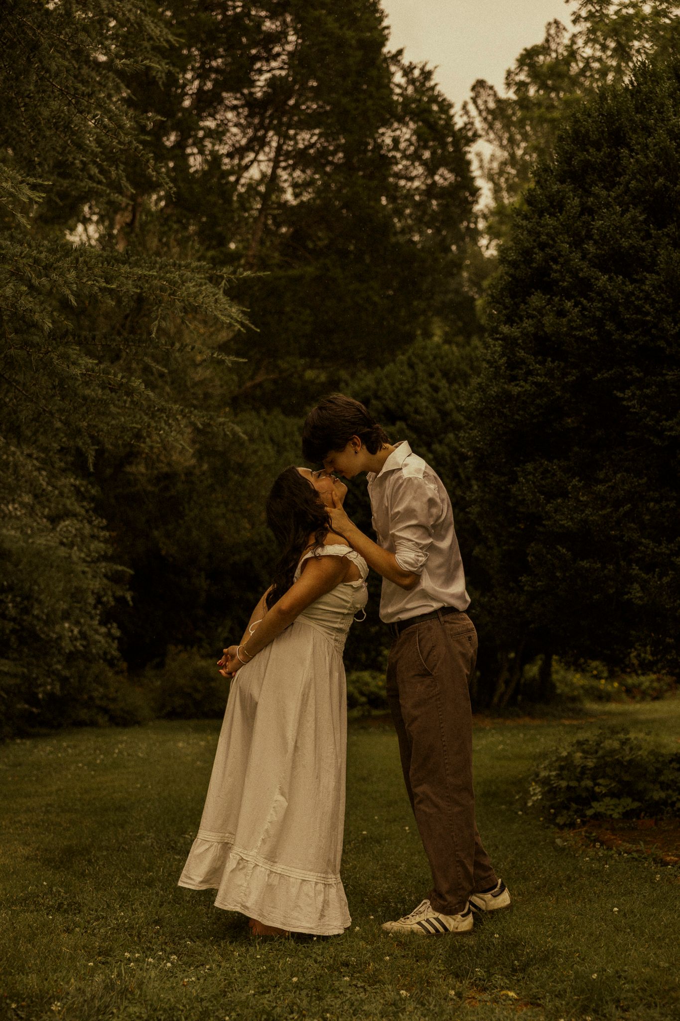 A couple shares a romantic kiss on a lush green lawn surrounded by tall trees in a warm, golden-toned outdoor setting.