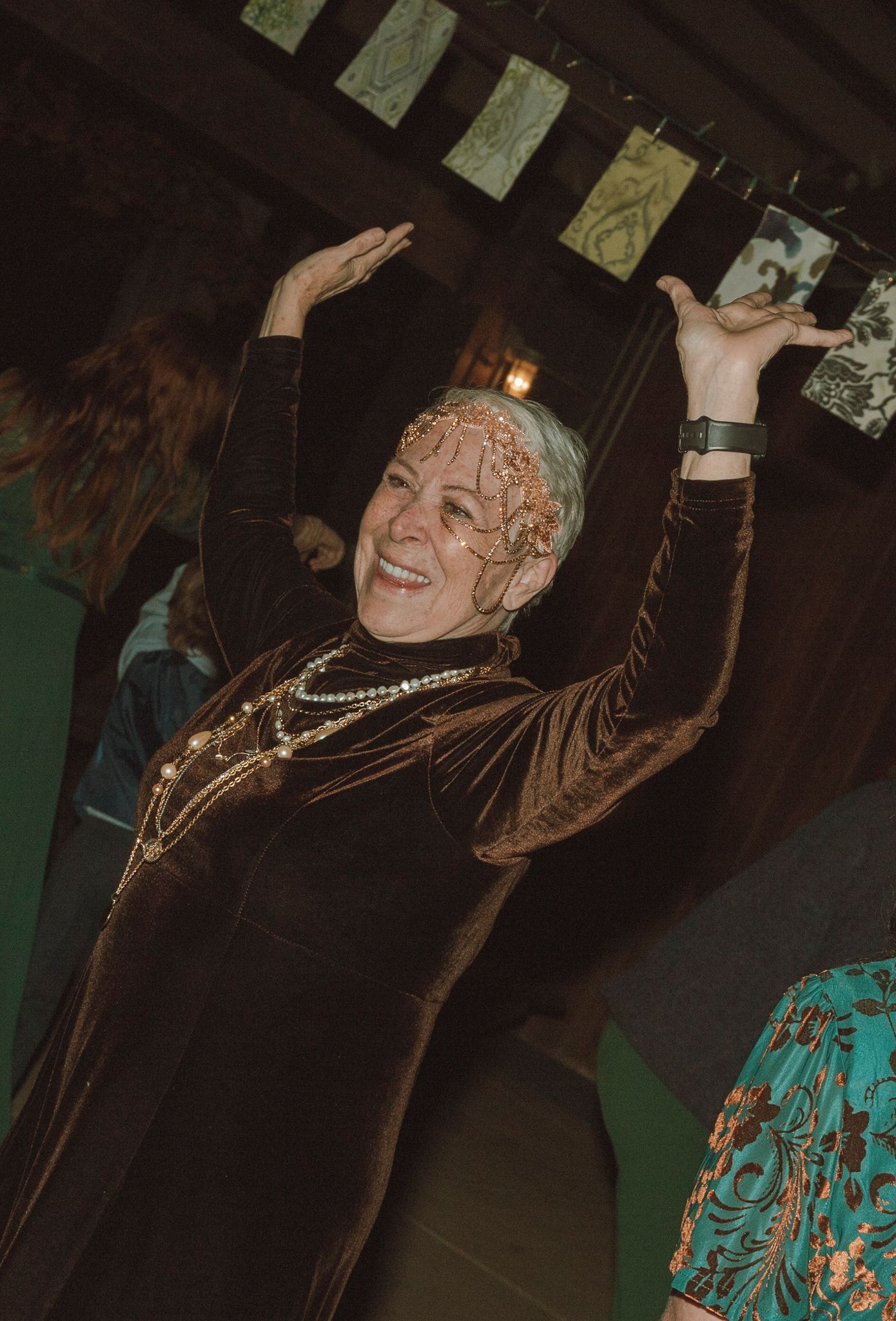 A person in a dark velvet jacket dances joyfully with raised arms under hanging decorations at an indoor celebration.