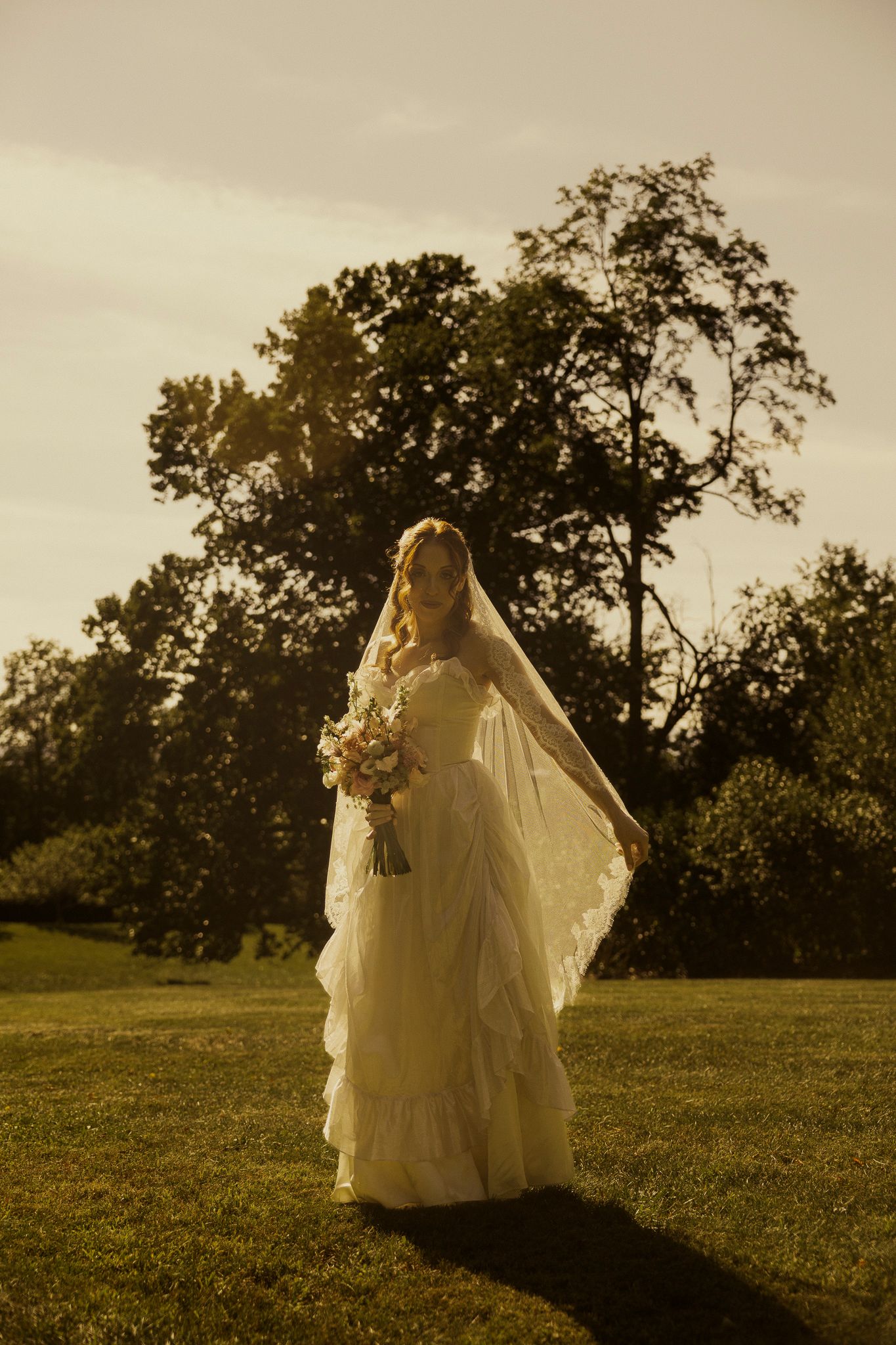 Bride in white ruffled gown and veil holds bouquet on green lawn at golden hour with trees behind.
