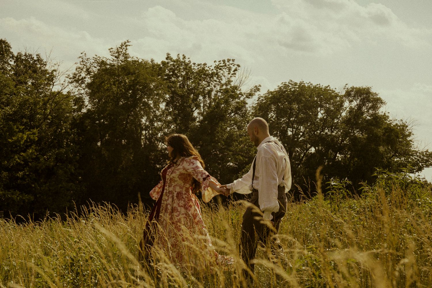Couple holding hands walking through tall golden grass field with lush green trees in background.