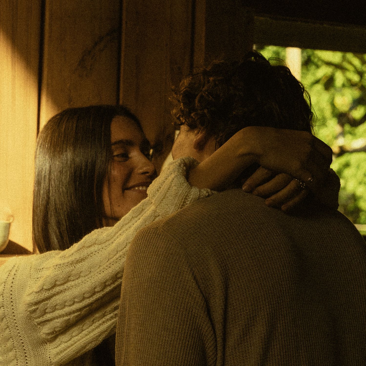 Two people sharing a warm embrace near a wooden wall, surrounded by soft natural light and green foliage.