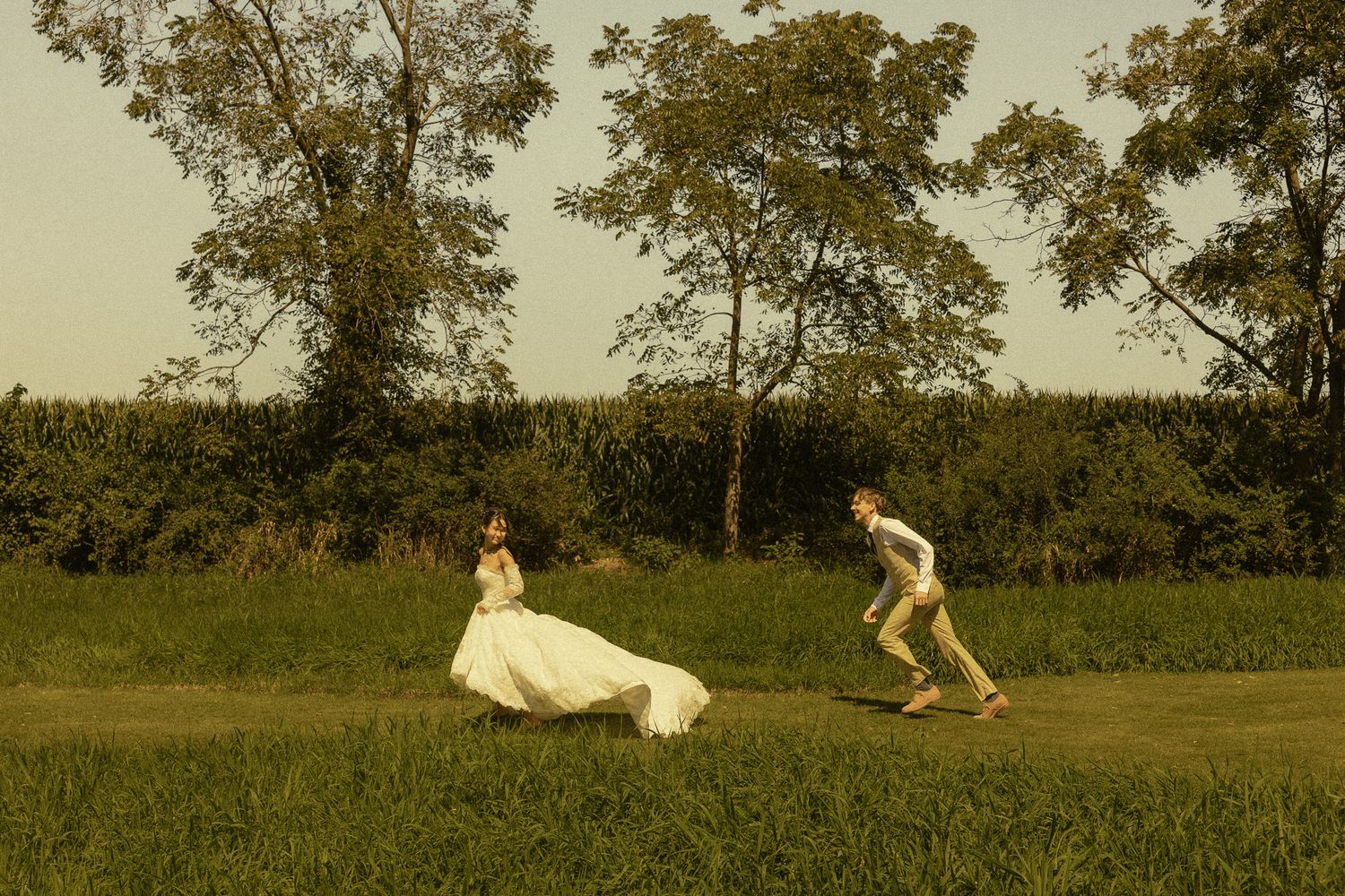 Bride in white gown and groom running joyfully through lush green fields at golden hour during outdoor wedding.